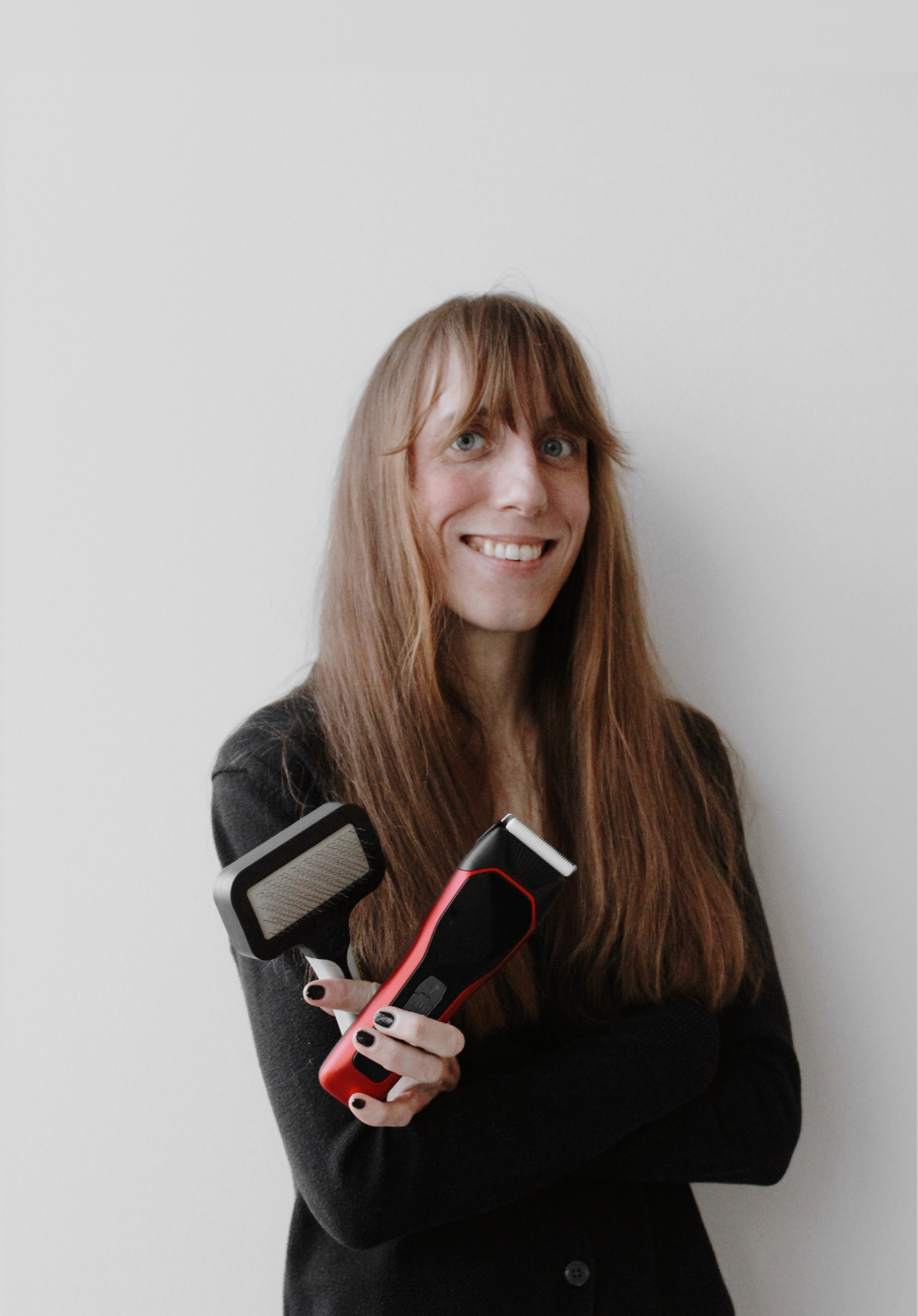 A woman with long reddish hair, smiling and holding an electric hair clipper with a brush attachment, standing against a plain white wall.