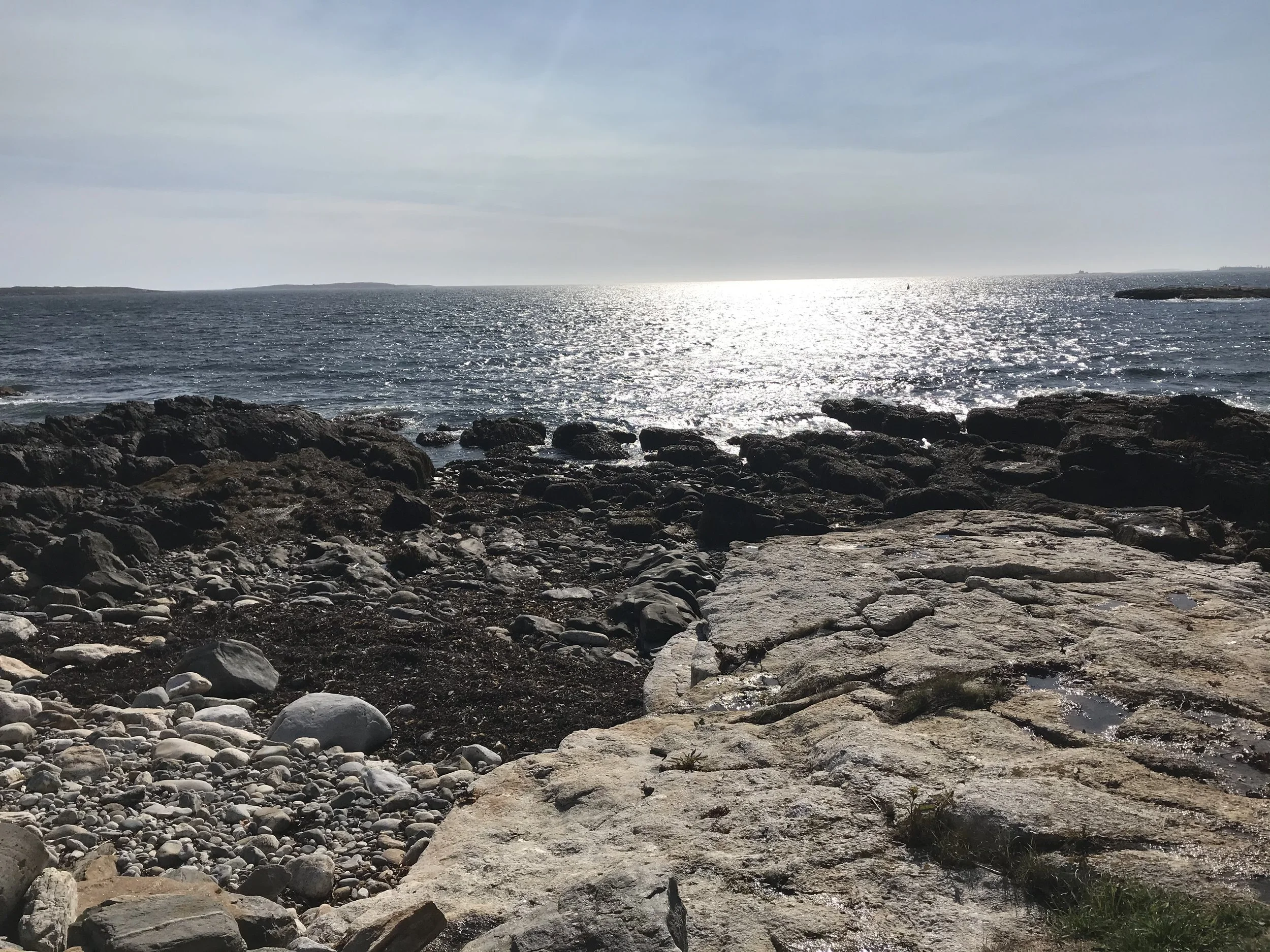 Rocky beach with large flat rocks in the foreground, smaller stones scattered along the shore, with the ocean extending into the horizon under a cloudy sky.