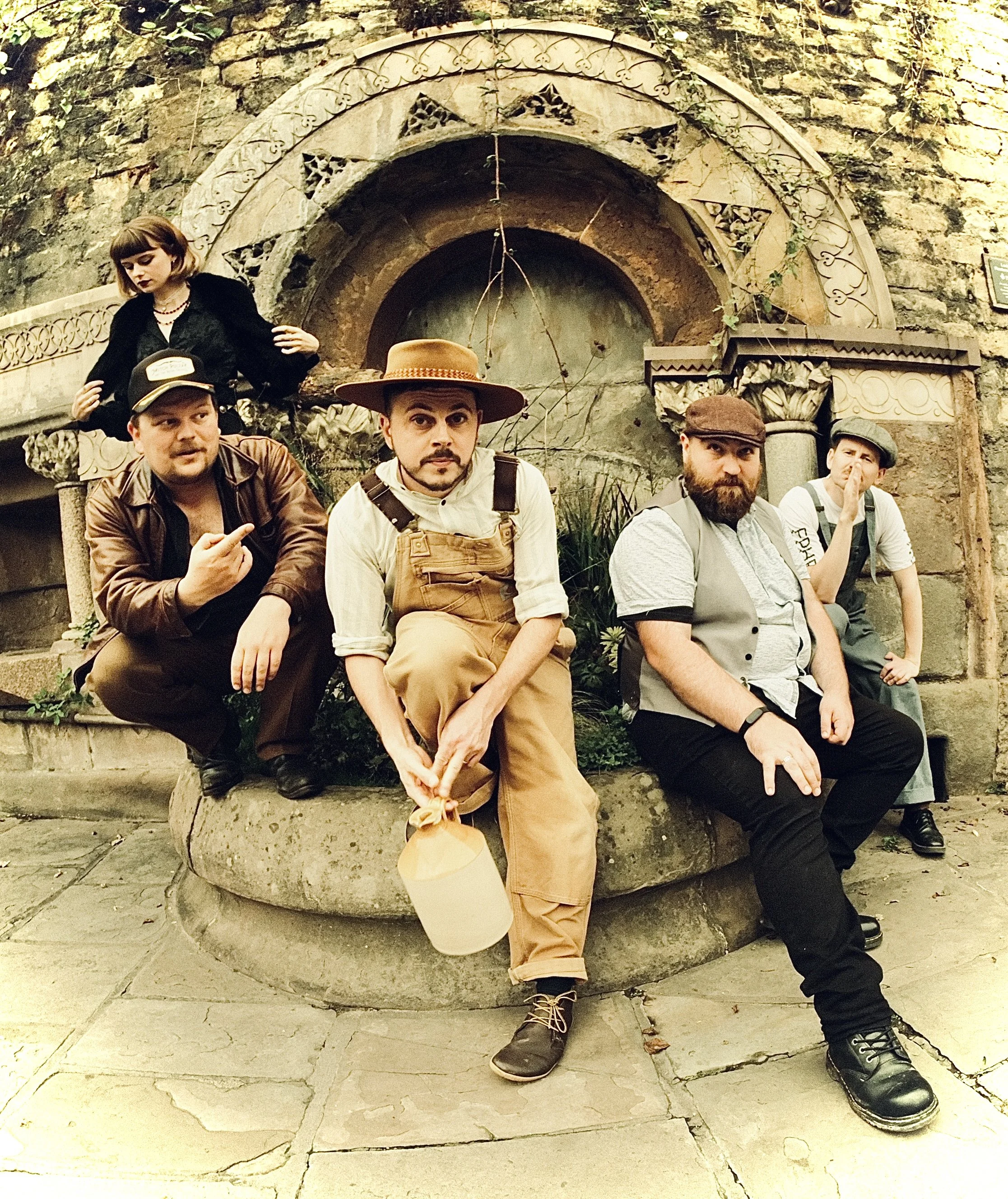 Five people dressed in vintage-style clothing sitting and crouching in front of an ornate, circular stone archway. One person holds a jug. Background features a stone structure with intricate carvings.