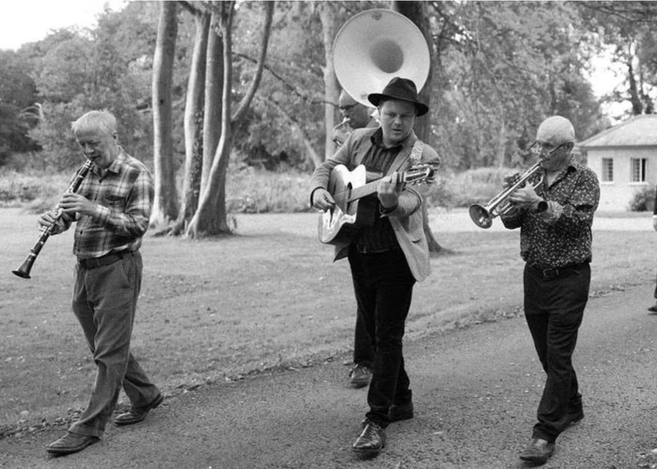 A black and white photo of three men walking outdoors while playing musical instruments. One man plays a clarinet, another plays a guitar, and the third plays a trumpet. They are dressed casually, and trees are visible in the background.