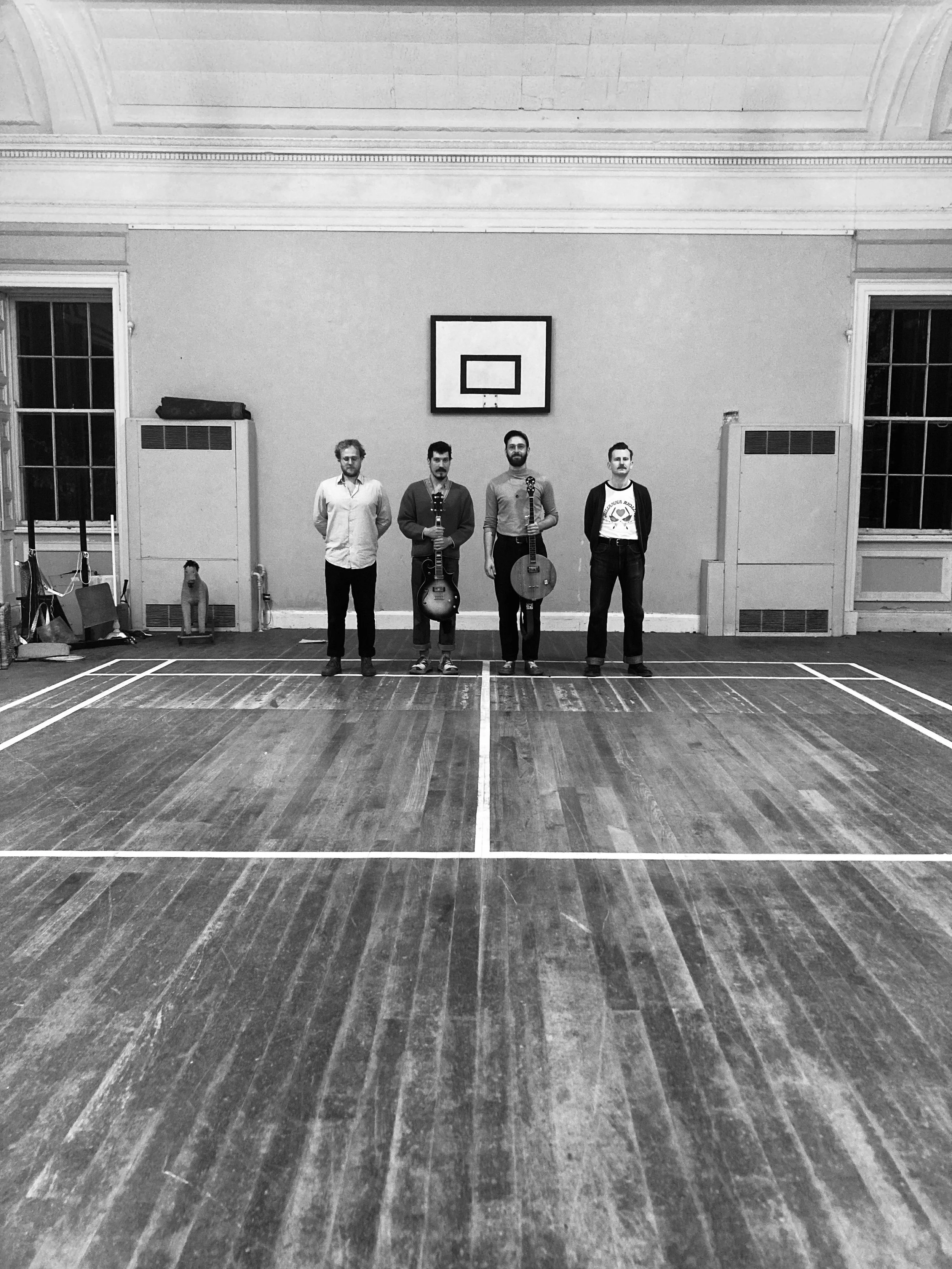 Four men standing on a wooden indoor court, holding guitars, with a basketball hoop on the wall behind them.