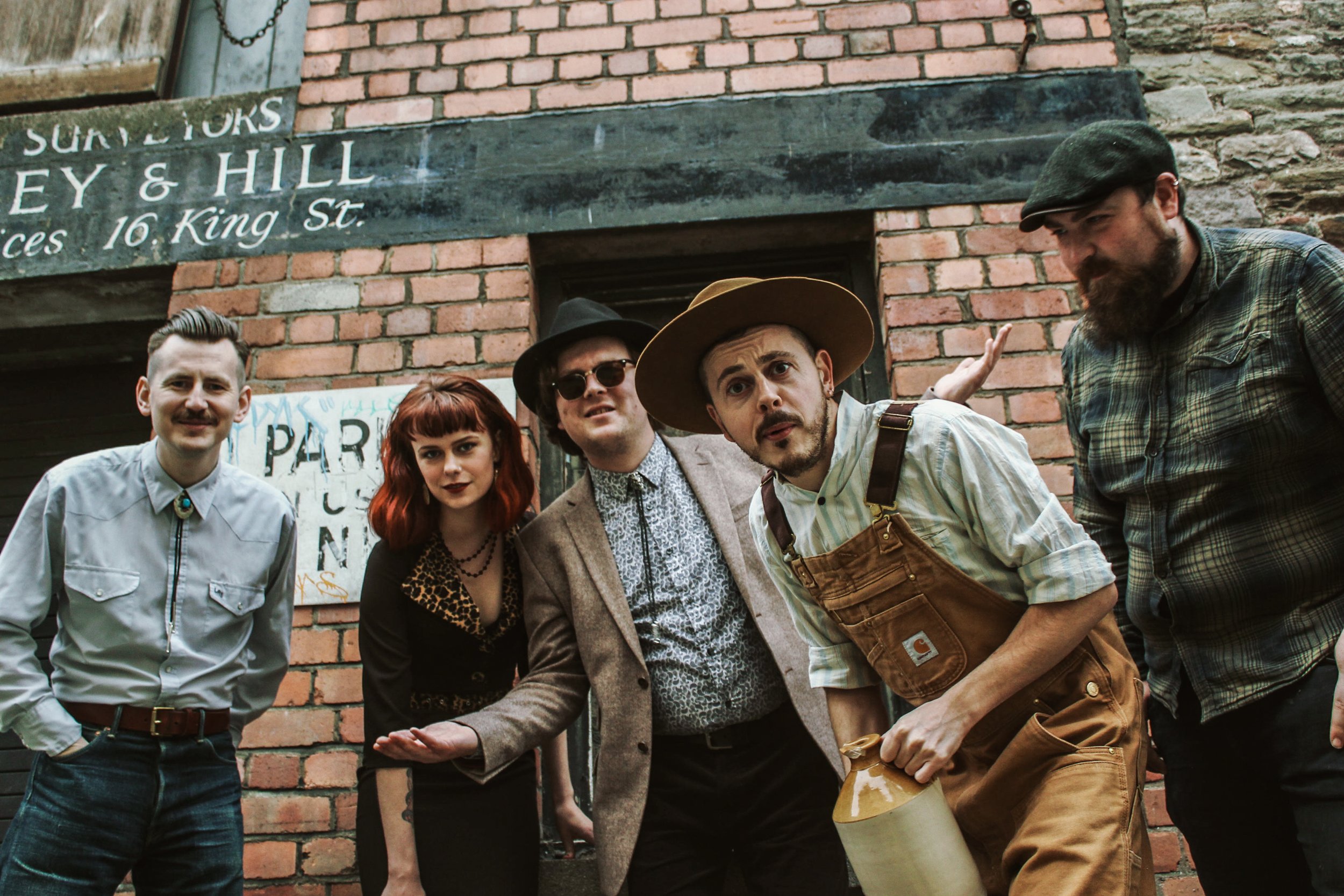 Group of five people posing in vintage clothing against a brick wall, with a mix of hats and casual wear.