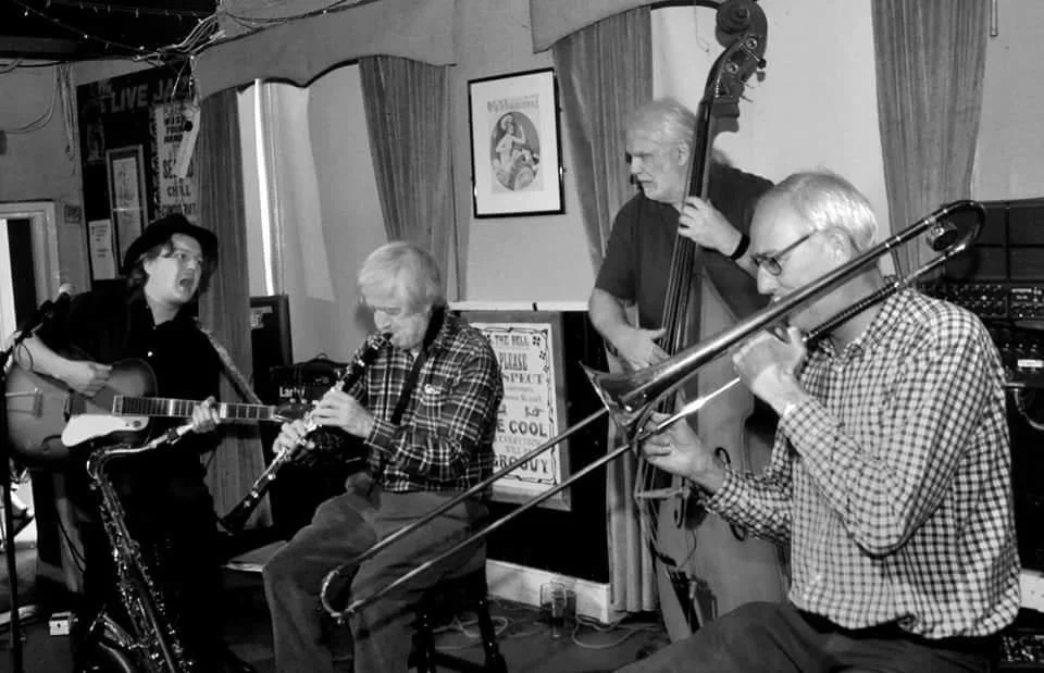 A black and white photo of four musicians performing in a small venue. The group includes a guitarist, a clarinetist, a double bassist, and a trombonist. The setting appears intimate, with music posters on the walls and curtains in the background.