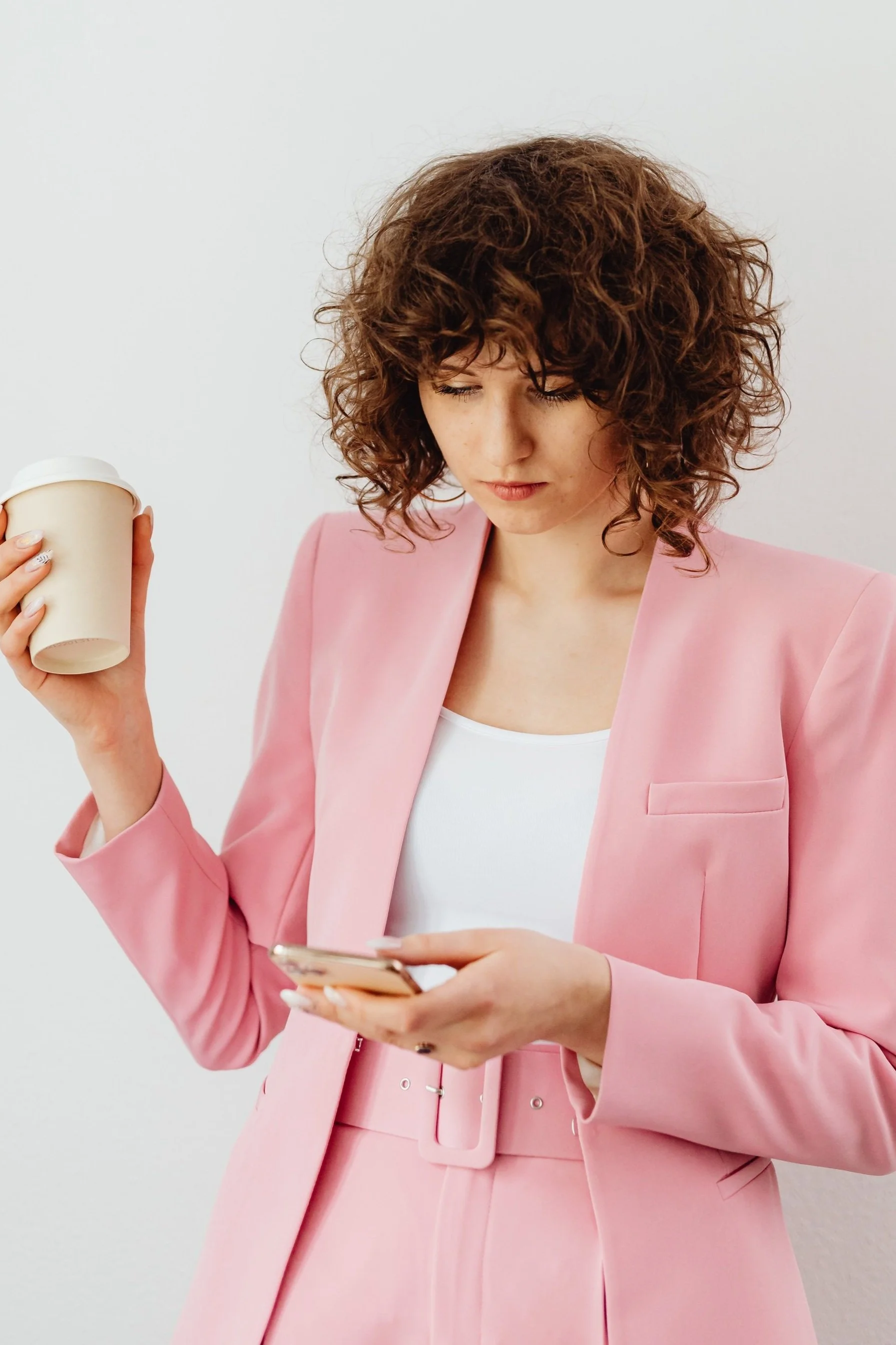 Woman in pink suit holding a coffee cup and a smartphone indoors.