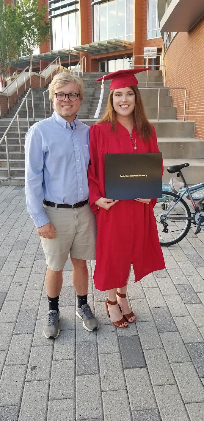 A young woman in a red graduation gown and cap holding a diploma from North Carolina State University, standing next to a young man outside a modern brick building on a paved area with stairs and a bicycle nearby.