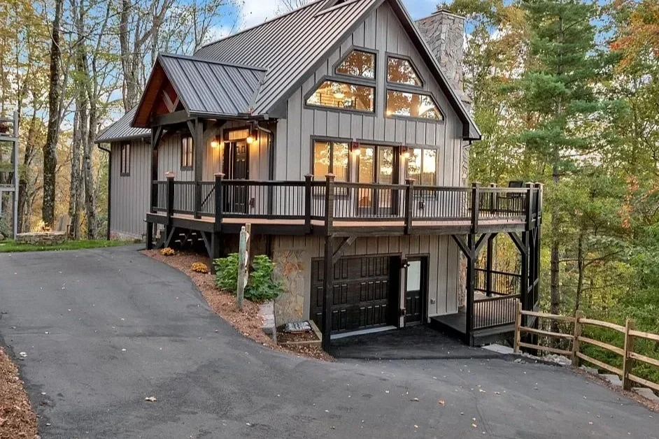 A gray two-story house with a black garage door, a deck, large windows, and surrounded by trees in autumn.