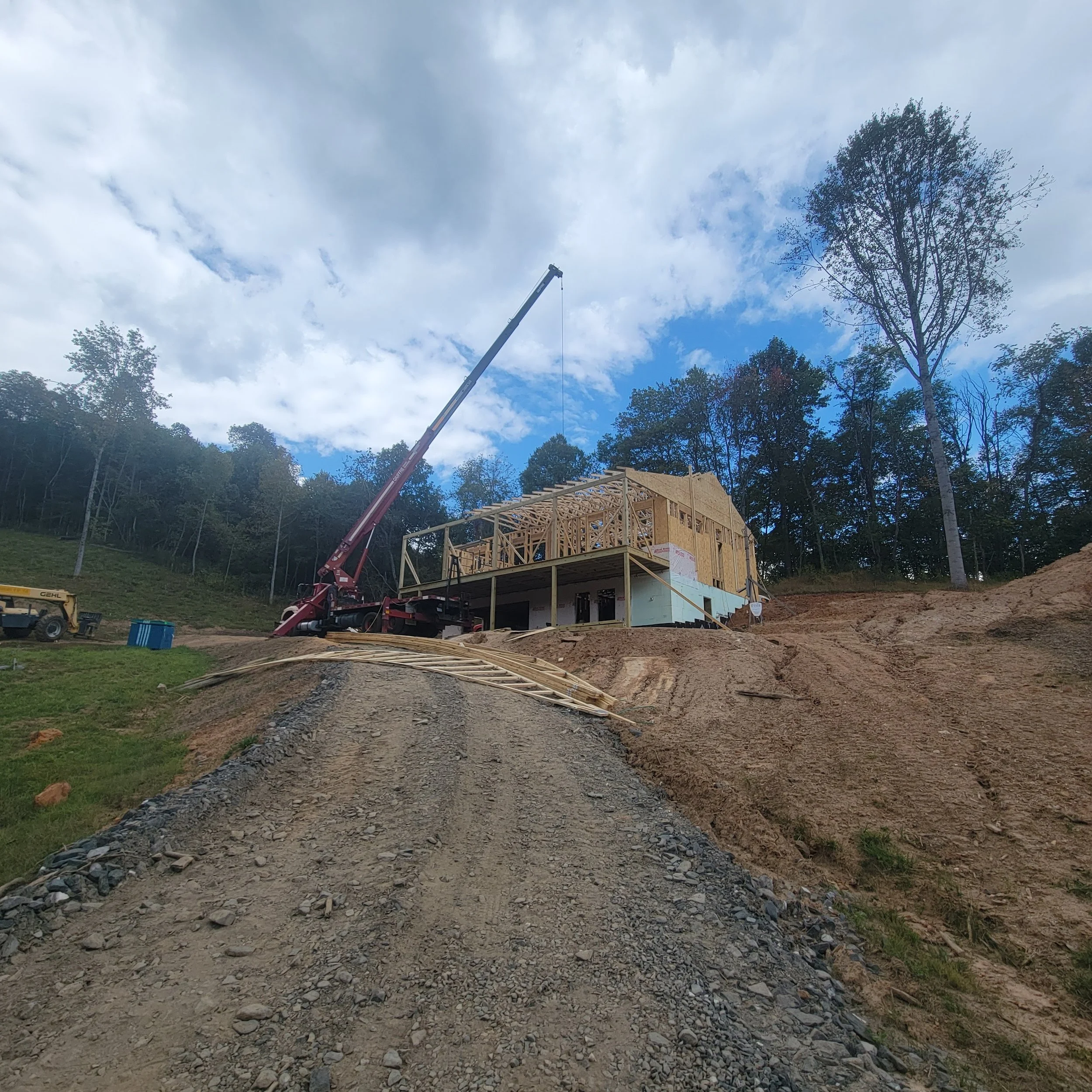 A house under construction on a hillside with a gravel driveway leading up to it, a crane lifting construction materials, and trees in the background under a partly cloudy sky.