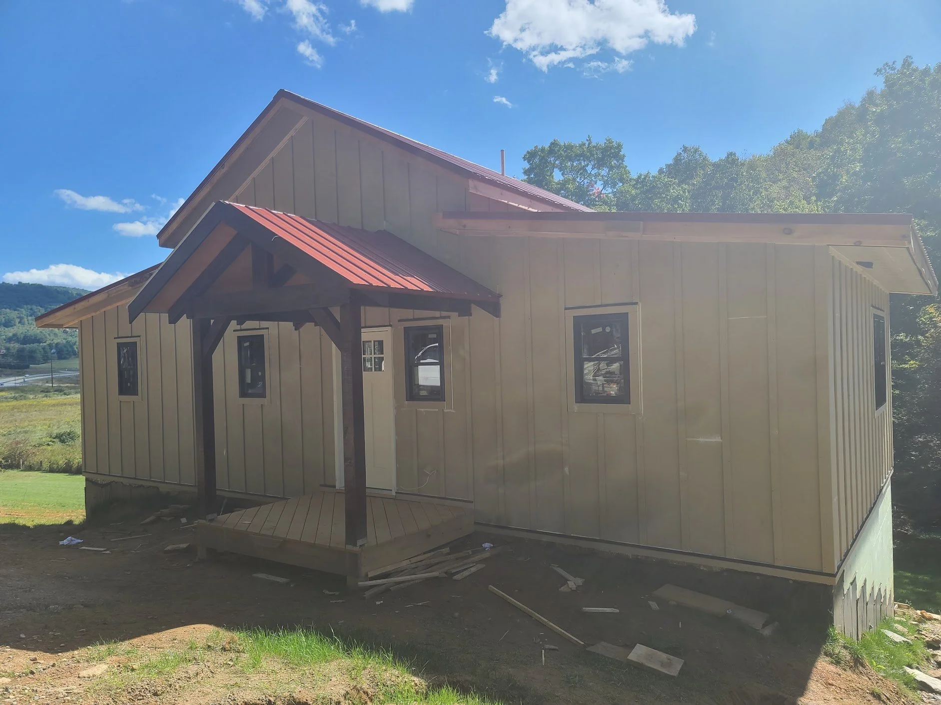 A small, beige house under construction with a red metal roof over the porch, set in a rural area with green fields and trees in the background.