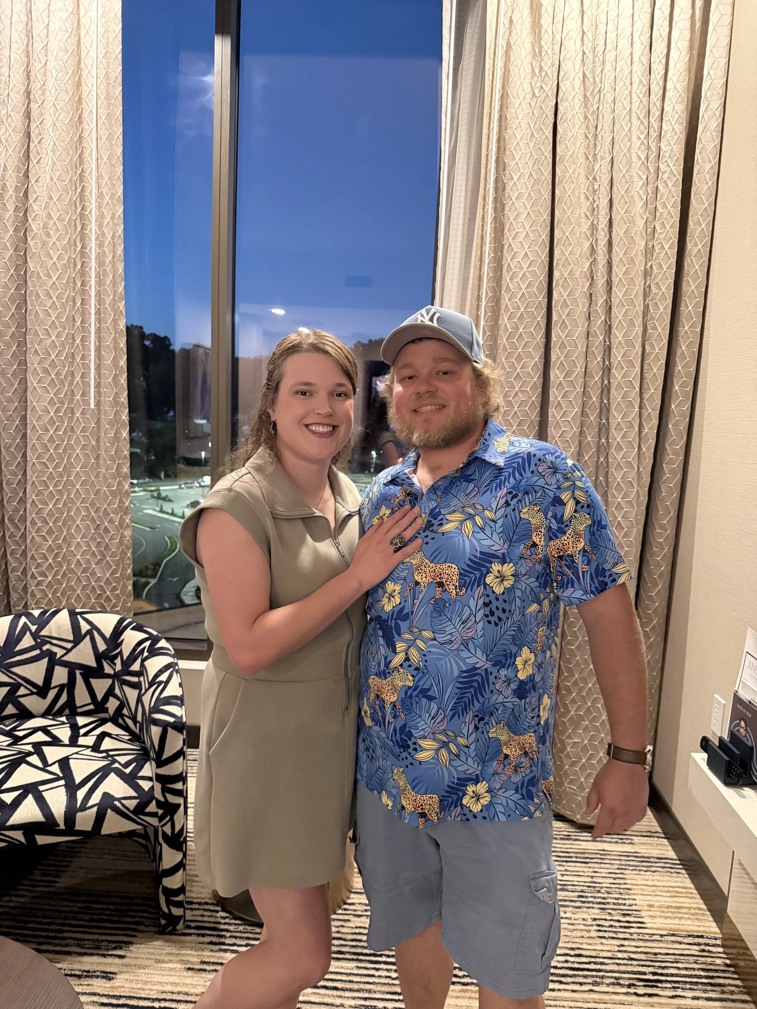 A man and woman smiling and posing together indoors, with a large window, patterned curtains, and outdoor scenery in the background.