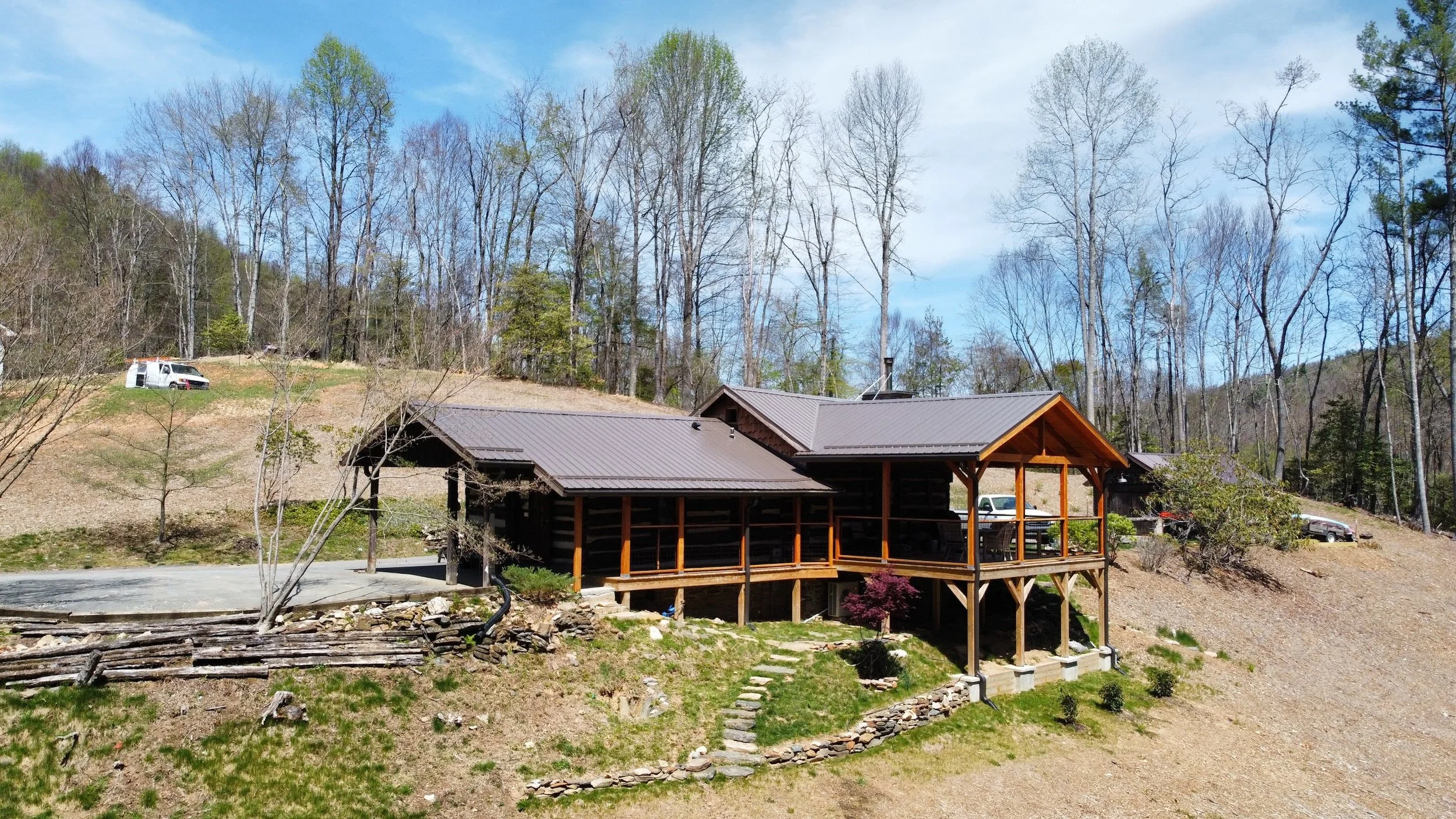A house with a covered porch and stairs, situated on a hillside with tall, leafless trees and a partly cloudy sky in the background.