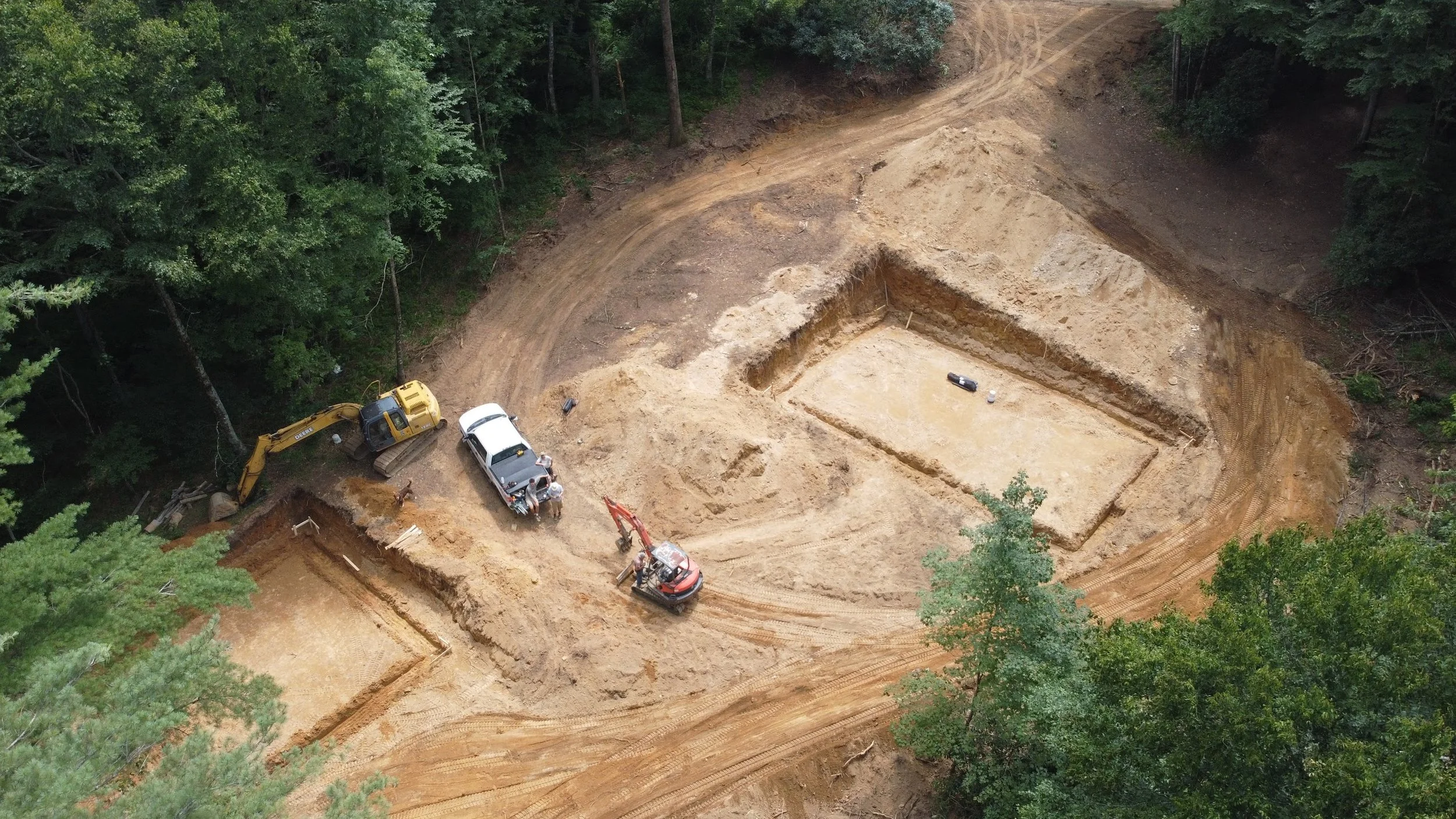 An aerial view of a construction site in a forested area with excavation equipment and vehicles, including a yellow excavator and a white pickup truck, preparing a large rectangular foundation in the dirt.