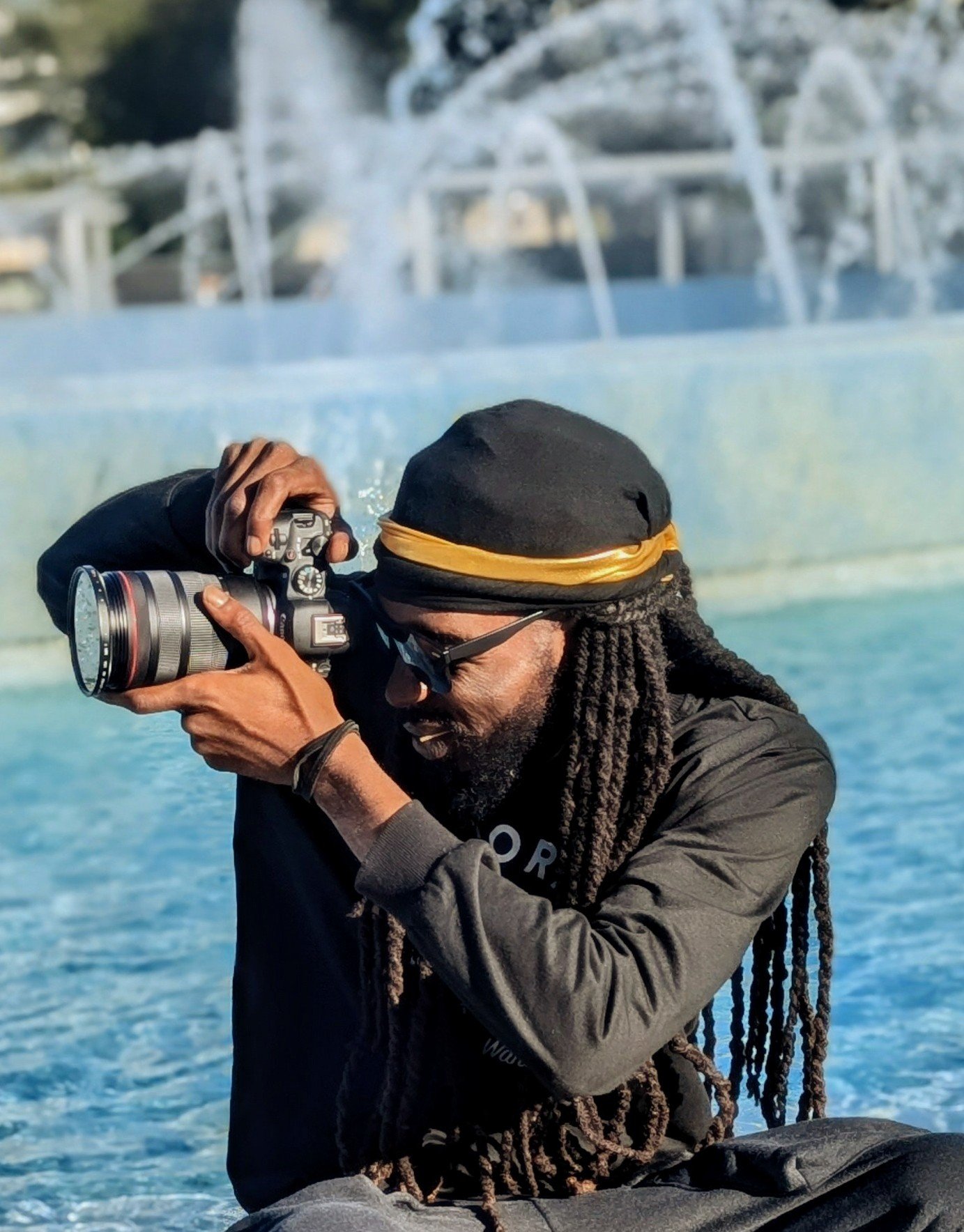 Dion Tyler of LocSmith Productions with long dreadlocks, wearing a black turban with a gold stripe, sunglasses, and a black crewneck, taking a photo with a camera near a water fountain with water features in the background.