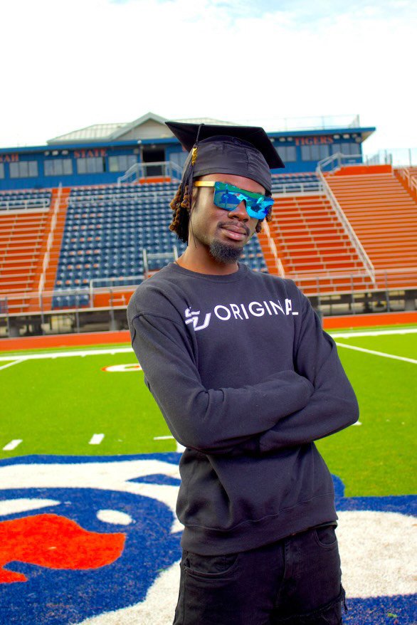 DJ MJ Da Kidd wearing a graduation cap and sunglasses stands with arms crossed on a sports field, with empty stadium bleachers in the background.