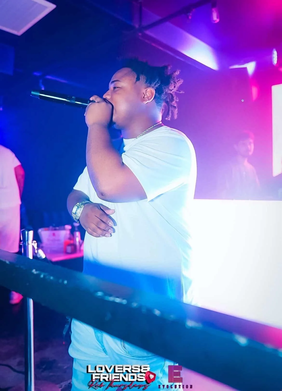 Party With Brice with dreadlocks holding a microphone to his mouth, singing at a karaoke or singing event. He is wearing a white t-shirt, a wristwatch, a chain, and appears to be in a club or lounge with purple and pink lighting.