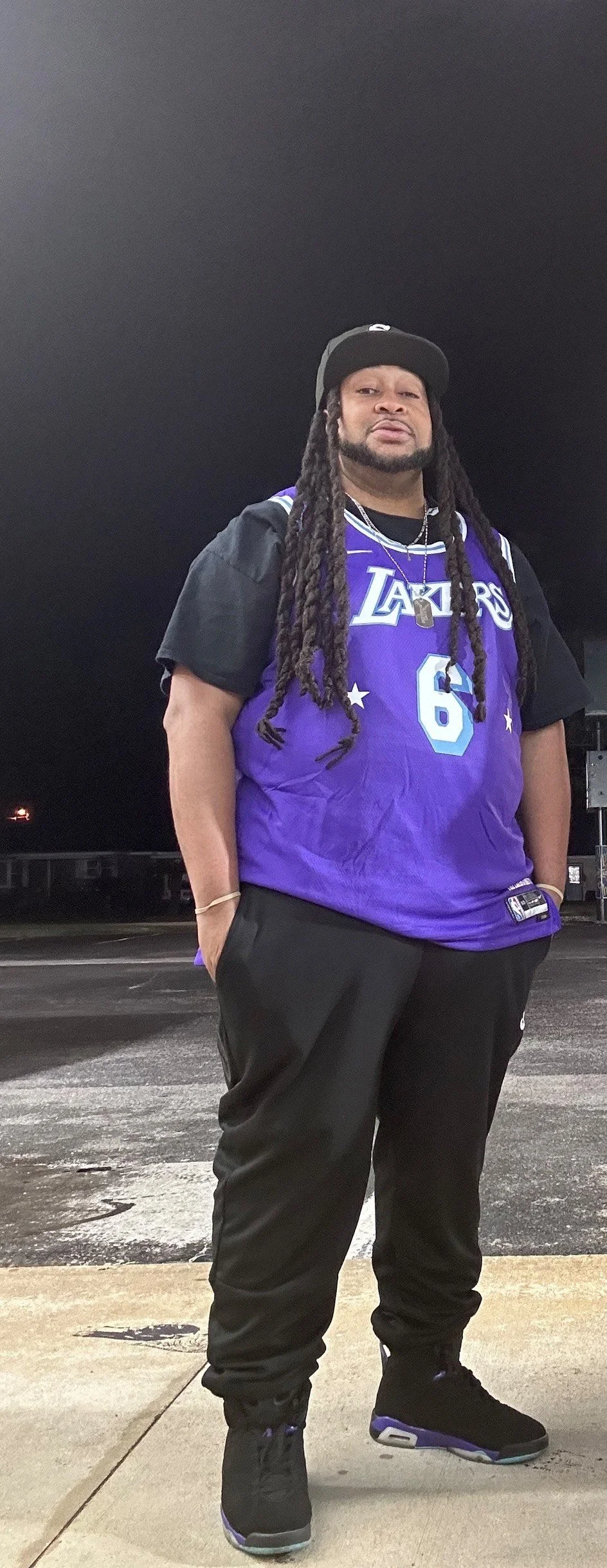 DJ Vnyce outdoors at night, wearing a purple Los Angeles Lakers basketball jersey with the number 6, black pants, black sneakers with purple and gray accents, a black cap, and necklaces, with his hands in his pockets.