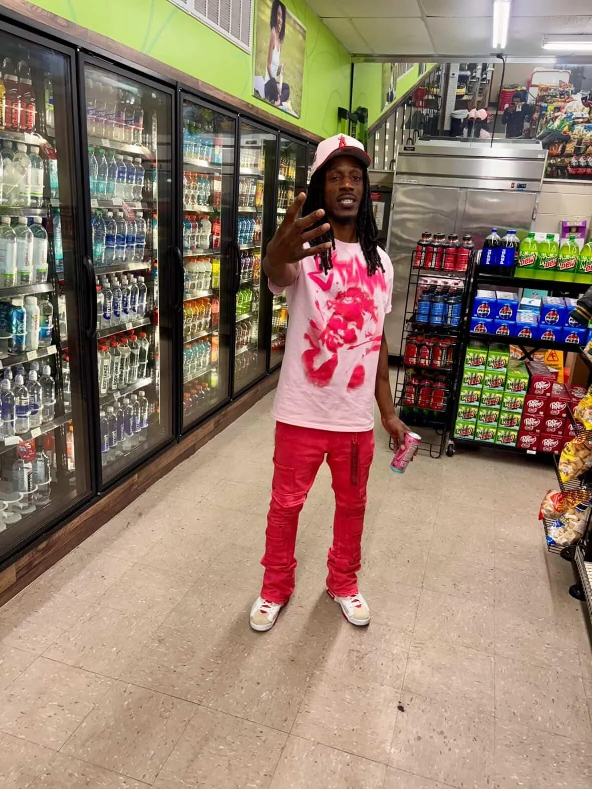 DJ Pawn in a pink t-shirt, red pants, and a red and white cap standing in a grocery store aisle, holding a drink, with grocery shelves and refrigerated drinks in the background.