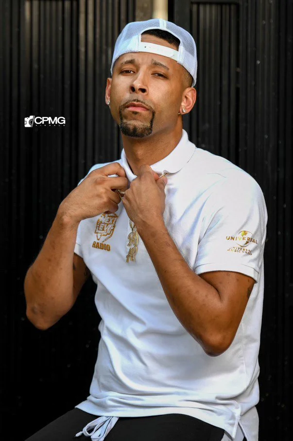 DJ AP Productions, wearing a white cap backward and a white t-shirt, pulling at his shirt collar with a confident expression, standing against a black background.
