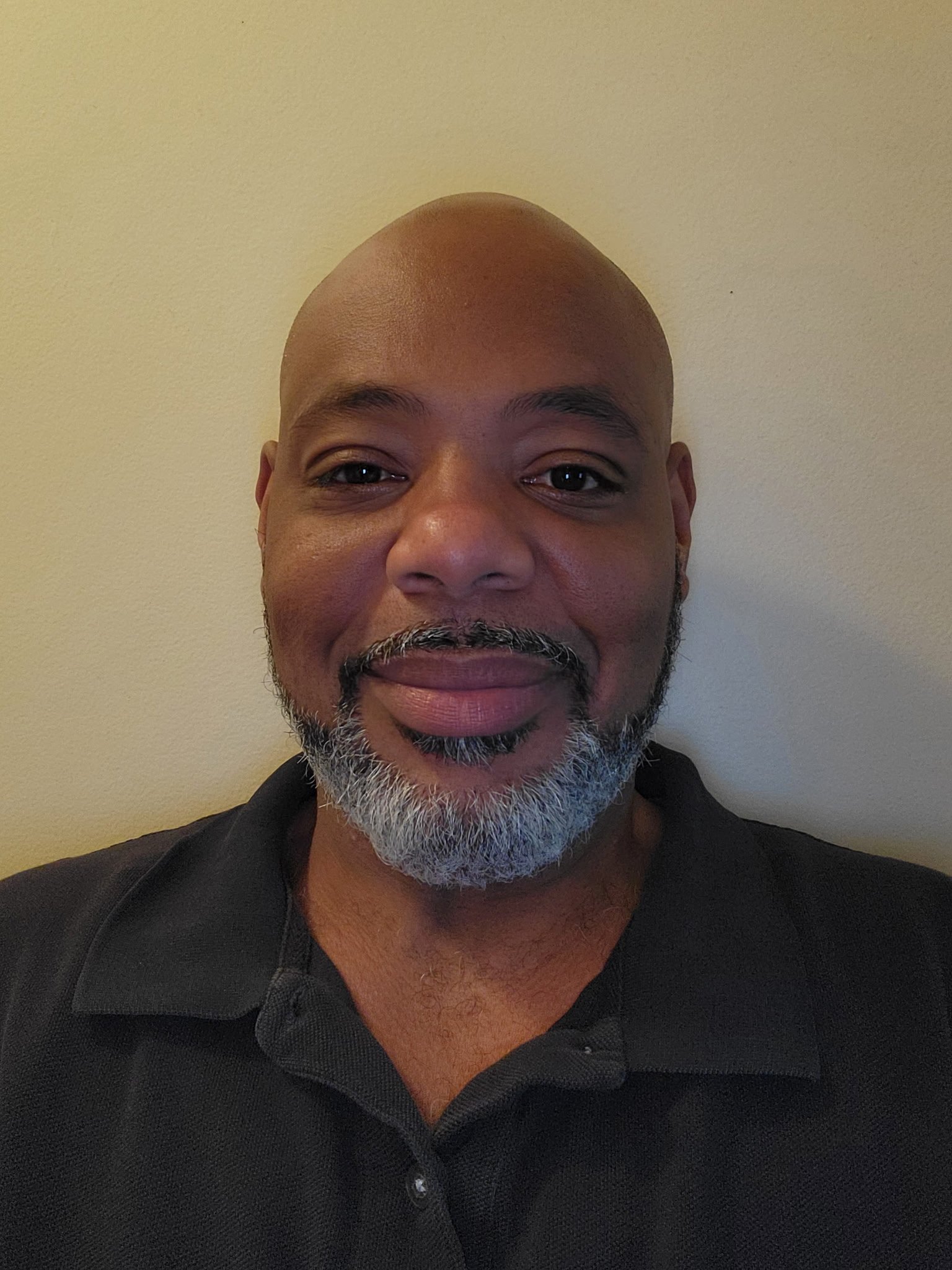 Close-up of a smiling middle-aged man with a shaved head, gray beard, wearing a black collared shirt, standing against a neutral beige background.