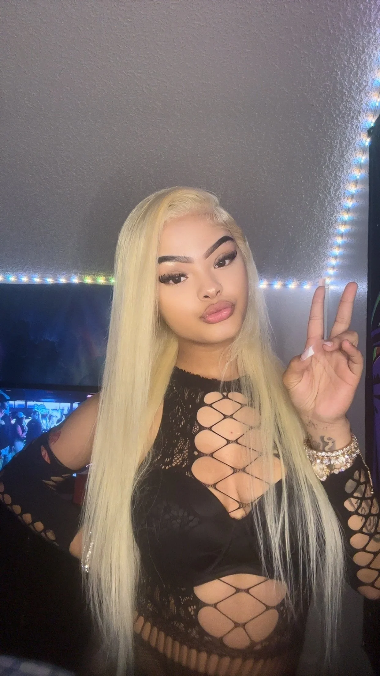 Madison Patterson with long blonde hair taking a selfie in a room with colorful LED lighting, wearing black fishnet clothing with cut-out details and jewelry.