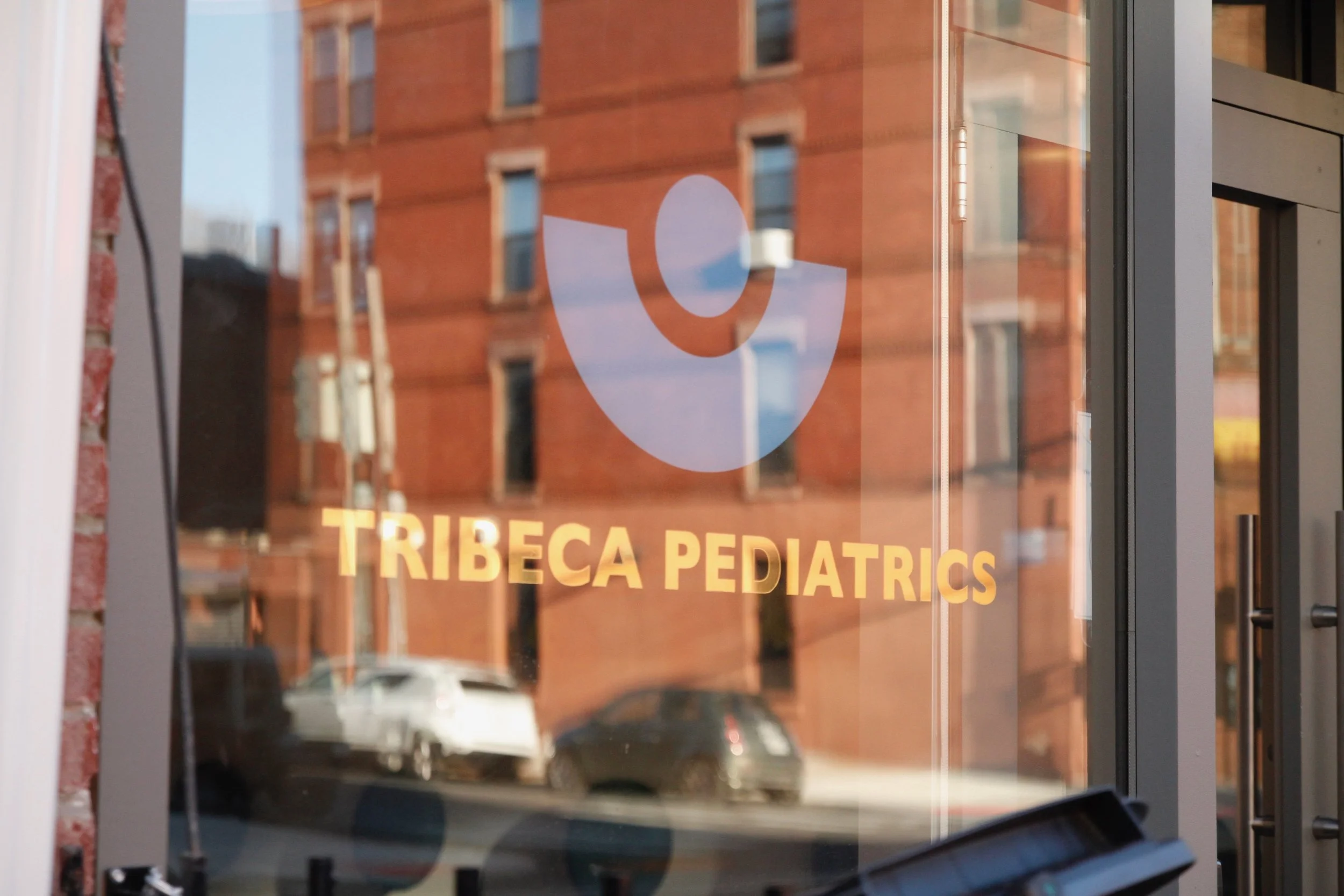 Glass storefront with Tribeca Pediatrics logo and bold gold lettering, reflecting historic red-brick architecture.