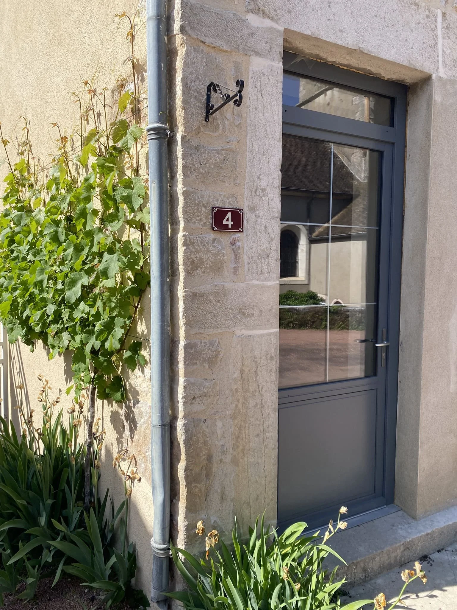 A close-up of a house entrance with a gray door, stone wall surrounding it, and a window above the door. A metal drainpipe runs along the left side, with green plants and flowers nearby. There is a house number 4 on a small plaque next to the door.