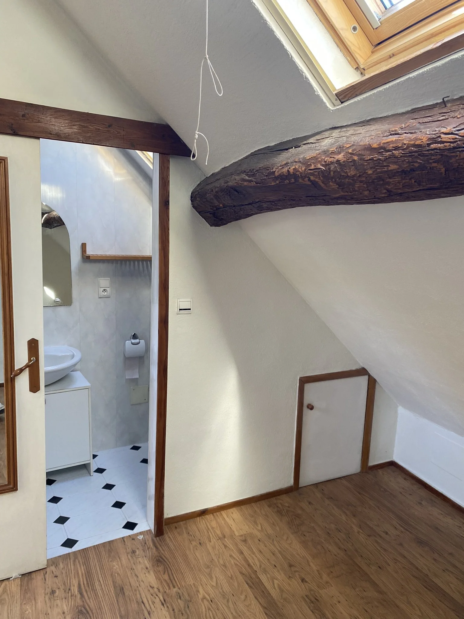View of a small bathroom with white tiled walls and black diamond-shaped accents, a white sink, and a mirror, seen through an open doorway from an attic space with wood flooring and a large dark wooden support beam.