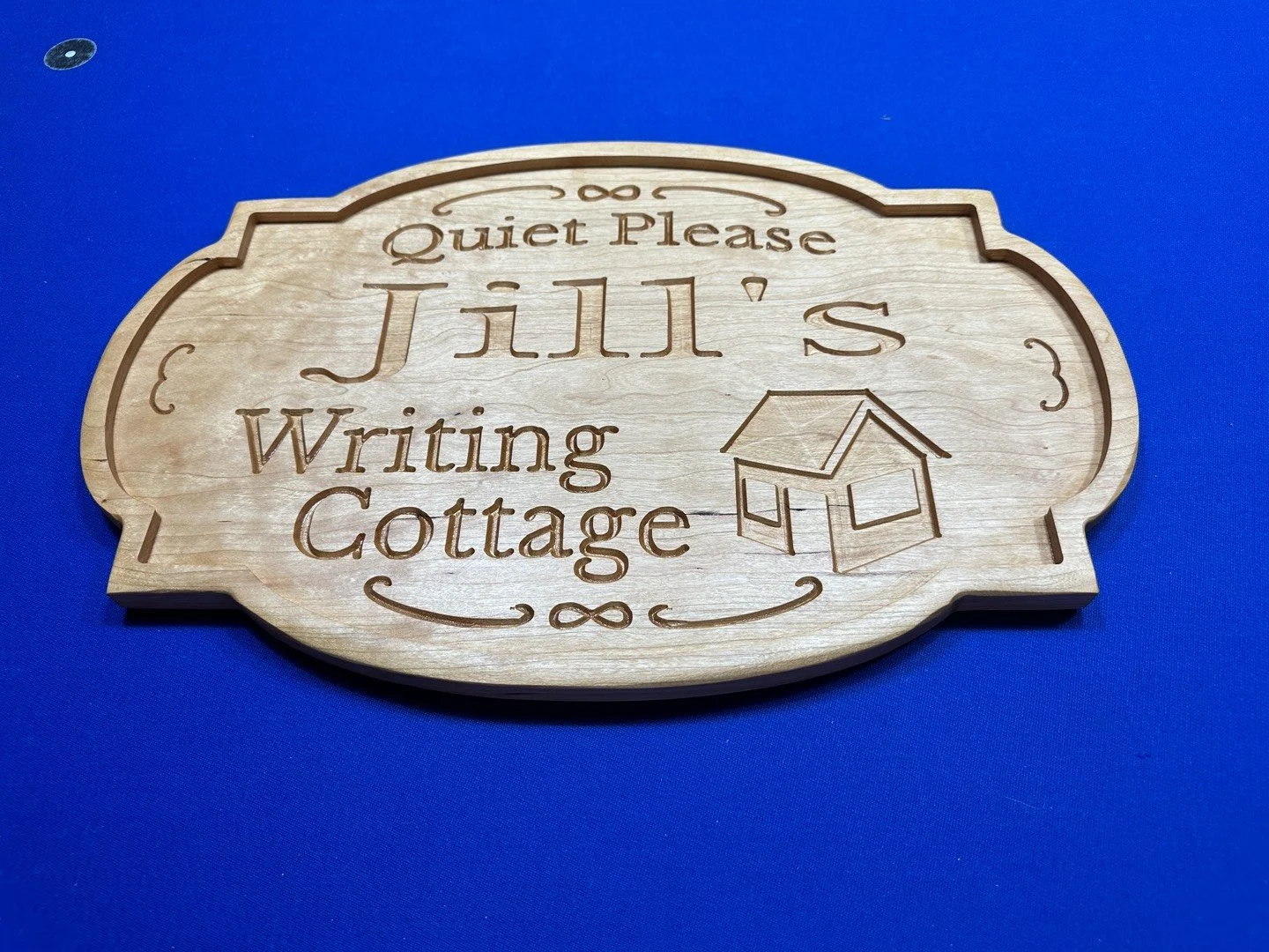 Wooden sign with engraved text reading "Quiet Please Jill's Writing Cottage" and a small illustration of a cottage, placed on a blue surface.