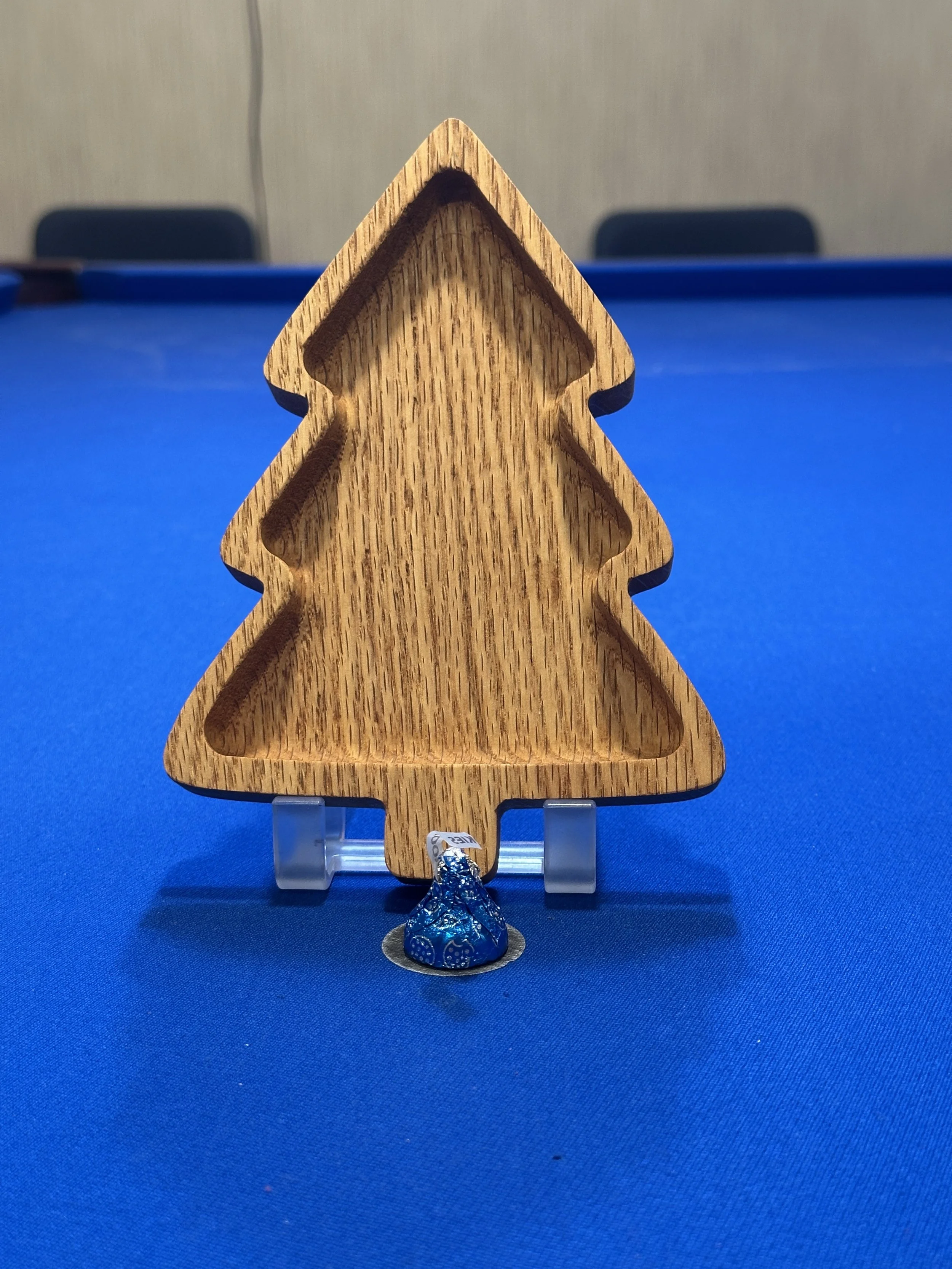 A wooden Christmas tree-shaped cutter placed on a blue surface, with a small wrapped candy wrapped in blue foil positioned at the base of the cutter.