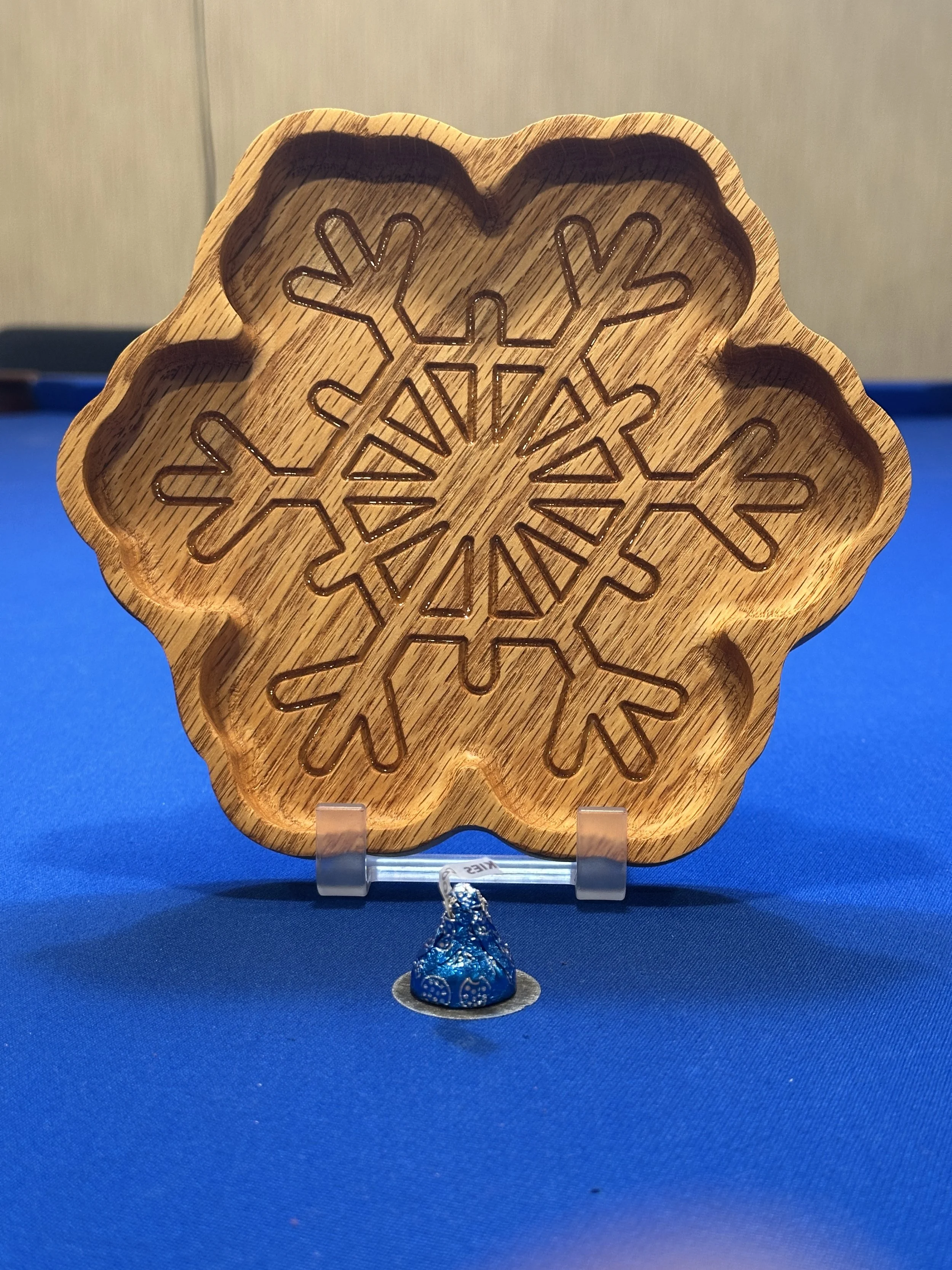 A wooden snowflake-shaped decoration with a cut-out snowflake design in the center, standing on a blue surface with a blue foil-wrapped chocolate piece in front.