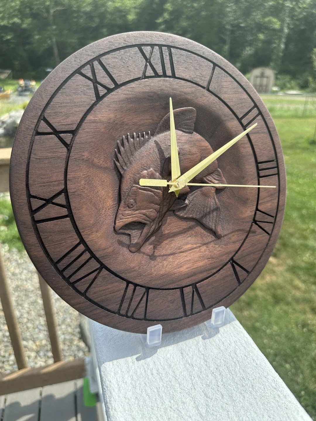 Wooden wall clock with Roman numerals and a carved fish design in the center, outdoors with blurred greenery and a lawn in the background.