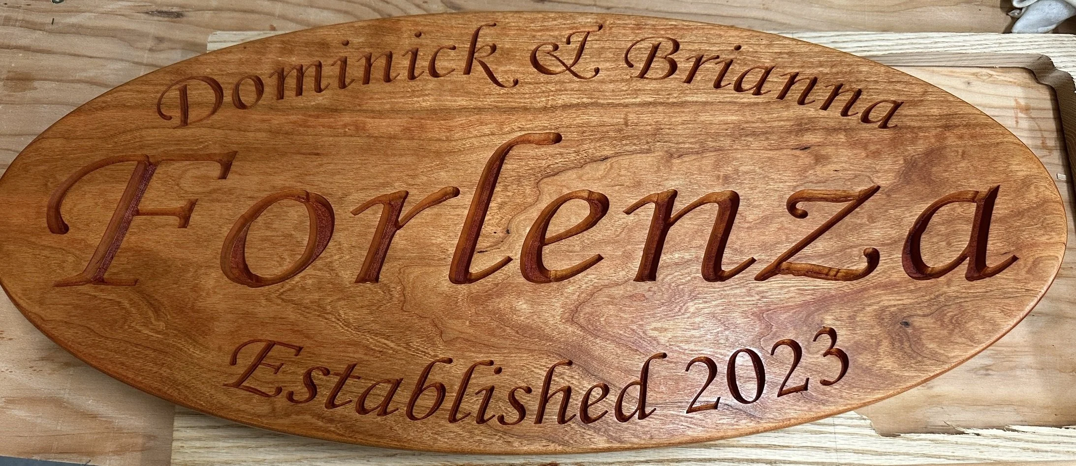 A wooden engraved sign with the text "Domnick & Briana Forlenza Established 2023" on an indoor wooden surface.