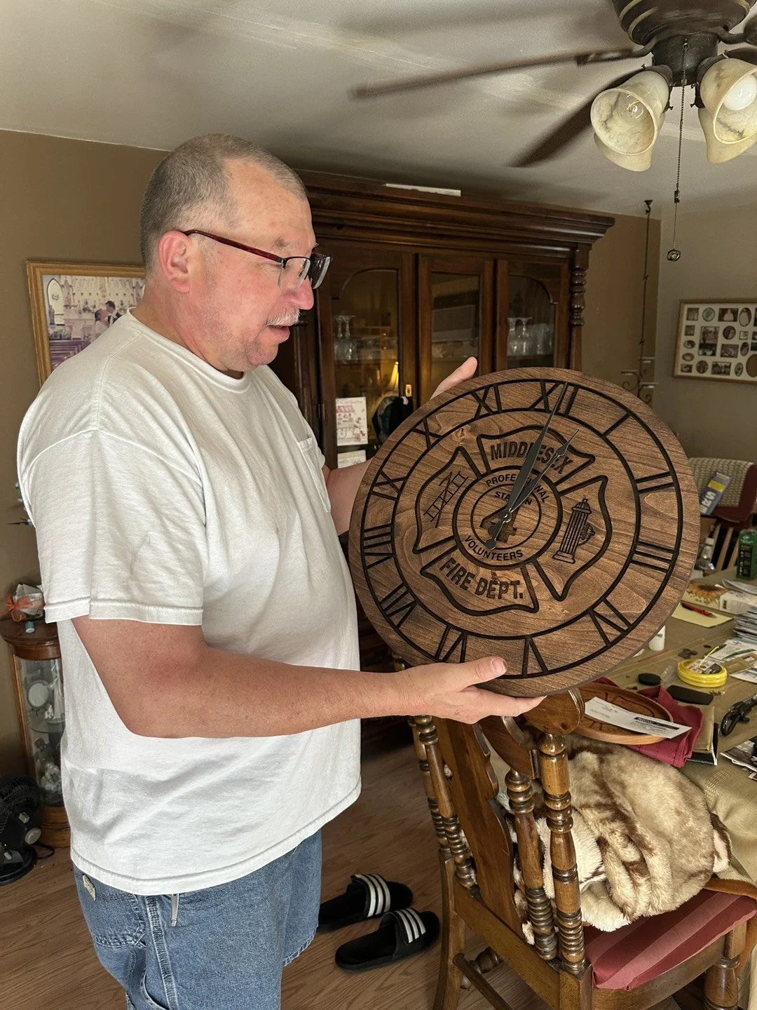 A man with glasses and a buzz cut hairstyle is holding a large wooden clock with Roman numerals. The clock has a design related to the Middlesex Volunteer Fire Department, including a firefighter badge, a water hydrant, and a ladder. The man is wearing a white t-shirt and jeans, standing in a room with wooden furniture, a ceiling fan, and various household items.