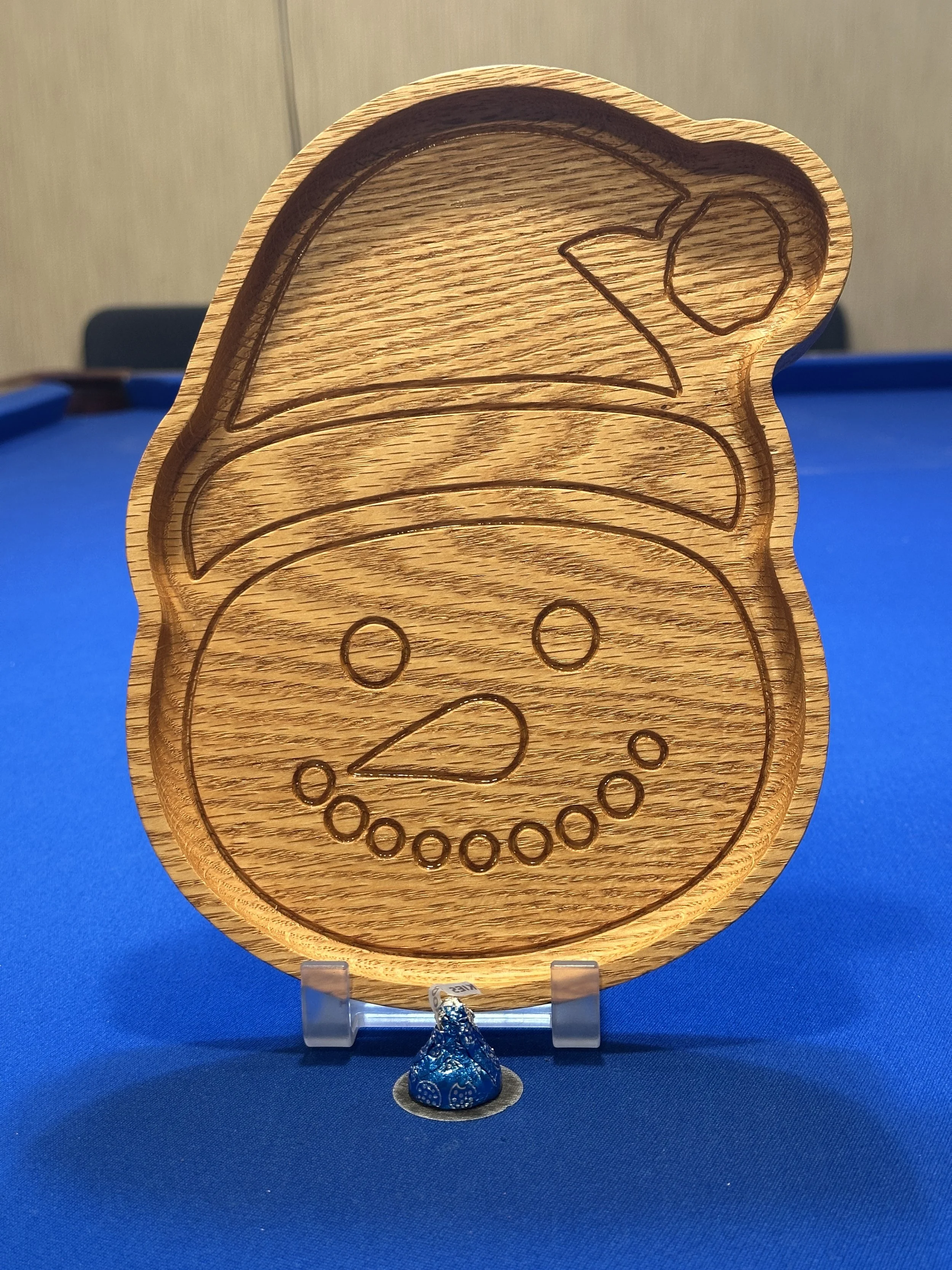 Wooden decorative item with a carved design of a face with a hat and a flower, placed on a blue surface with a small figurine at the base.