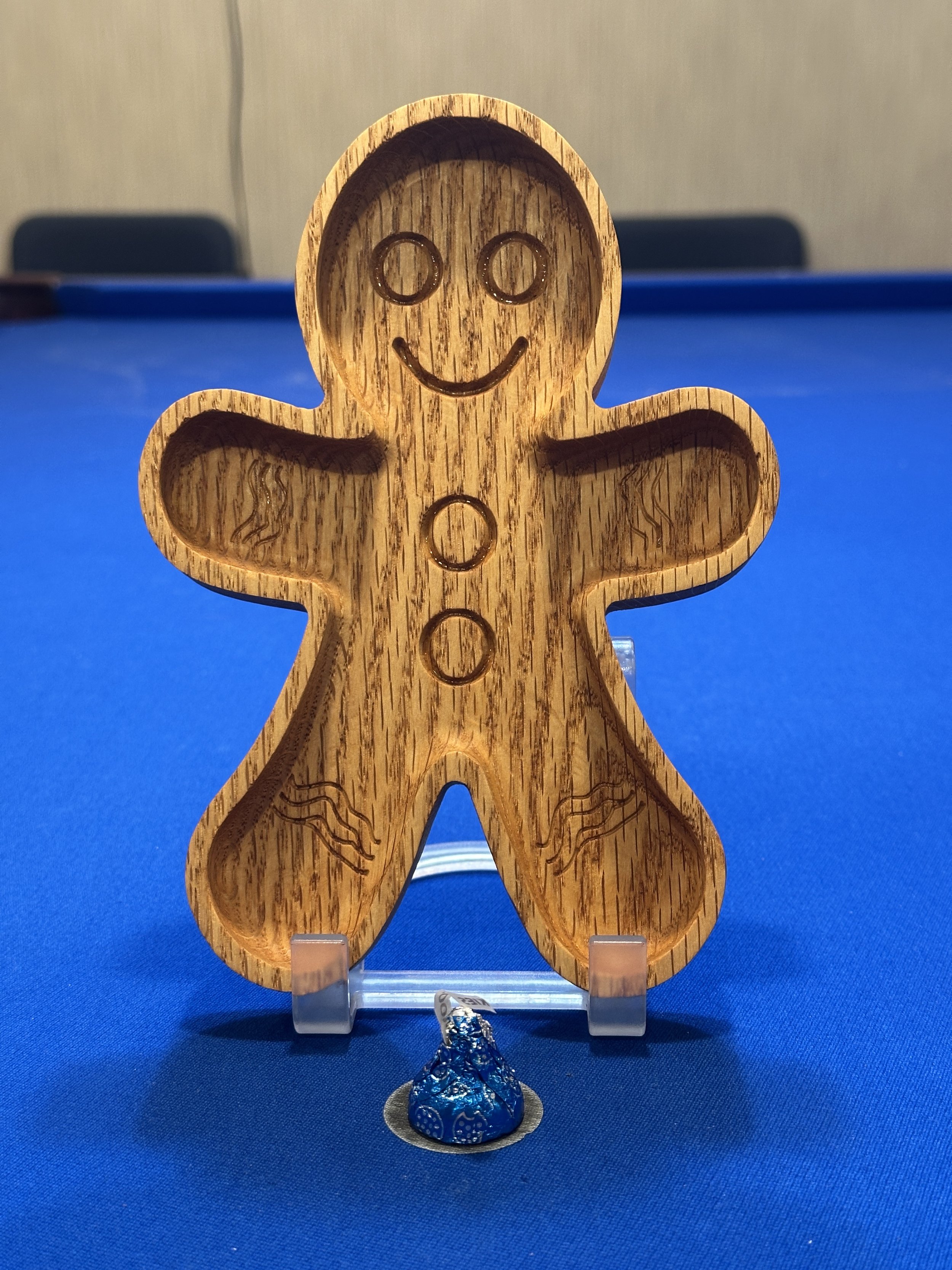 A gingerbread man-shaped wooden board with carved circles and a smiling face, sitting on a blue surface with a small wrapped chocolate treat at its base.