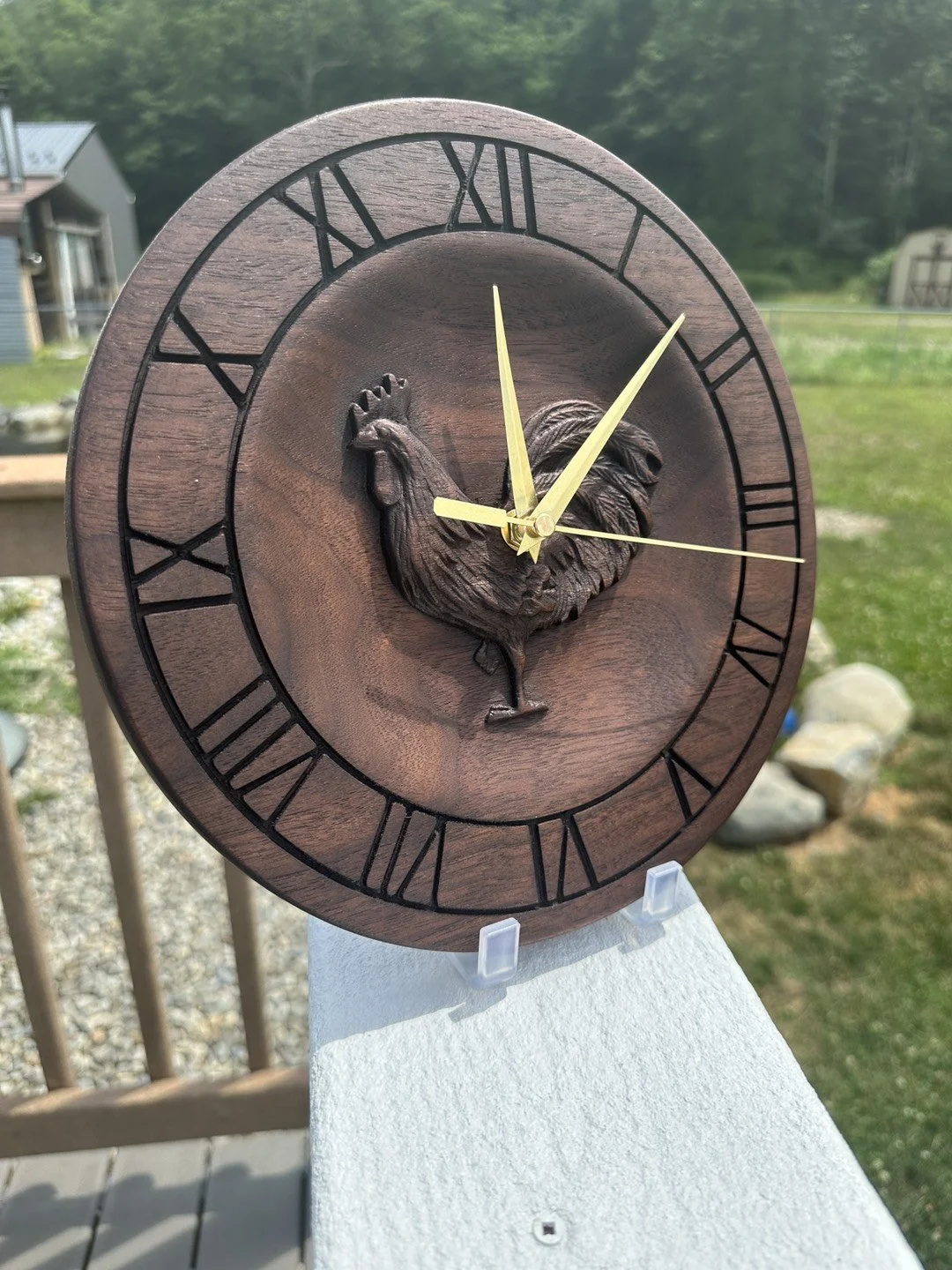 Round wooden wall clock with a carved rooster in the center, Roman numerals around the edge, gold hour and minute hands, and a gold second hand, set outdoors on a porch railing with a grassy yard and trees in the background.