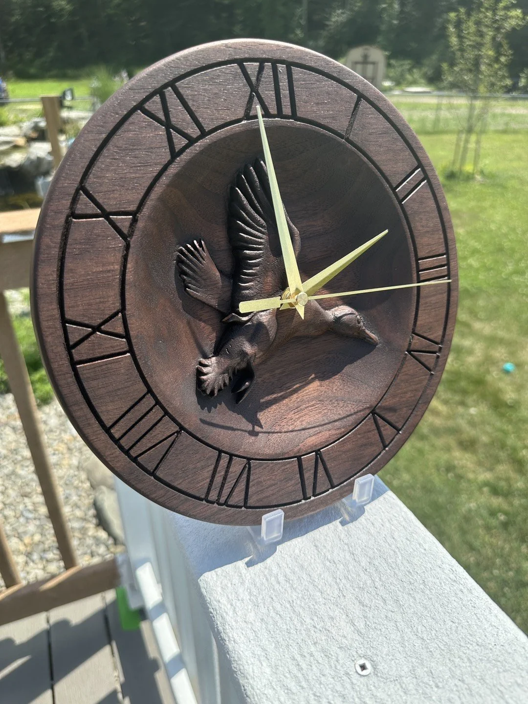 Wooden clock with Roman numerals and a carved hummingbird design in the center, outdoors with greenery in the background.