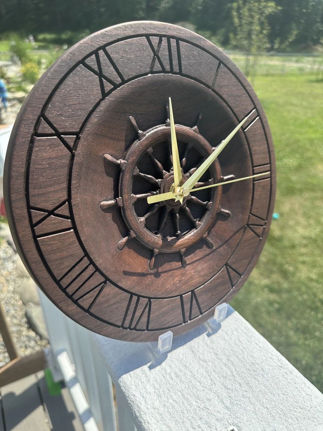 A round wooden wall clock with Roman numerals and a ship's wheel design in the center, showing the time as approximately 2:12.