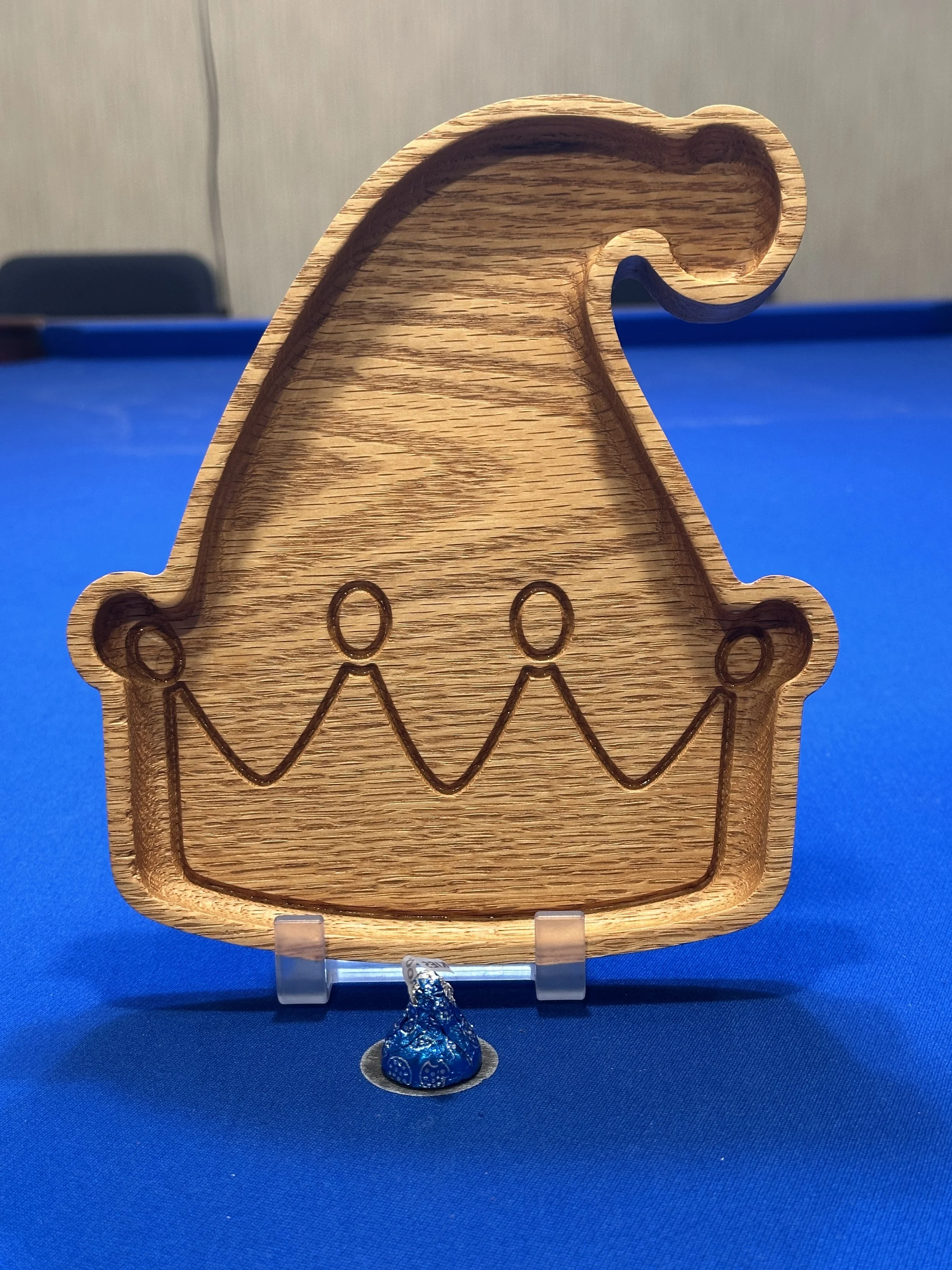 Wooden gingerbread cookie cutter in the shape of a gingerbread person, placed on a blue surface with a wrapped candy and a small circular support beneath it.