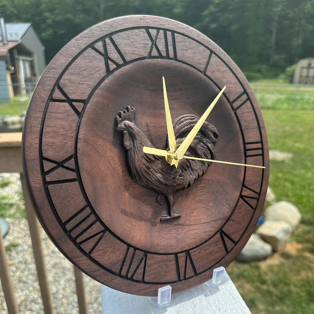 Round wooden wall clock with Roman numerals, featuring a carved rooster in the center, outdoors with grass and trees in the background.