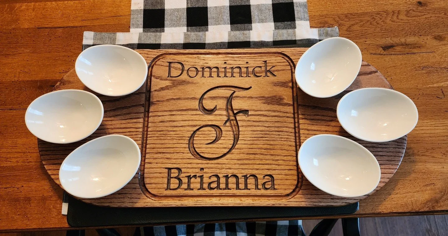 A wooden serving tray with the name 'Dominick Brianna' engraved in the center, surrounded by six empty white ceramic bowls on a wooden table with a black and white checkered cloth underneath.