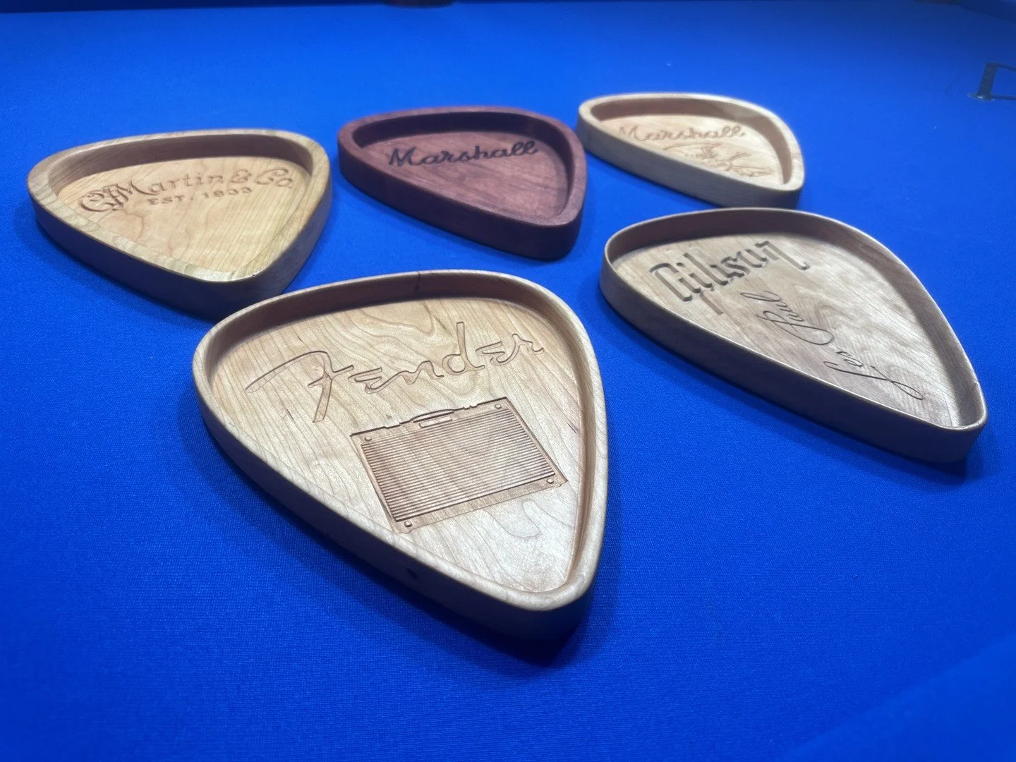 Five wooden guitar pick trays with engraved brand names and designs, placed on a blue surface.