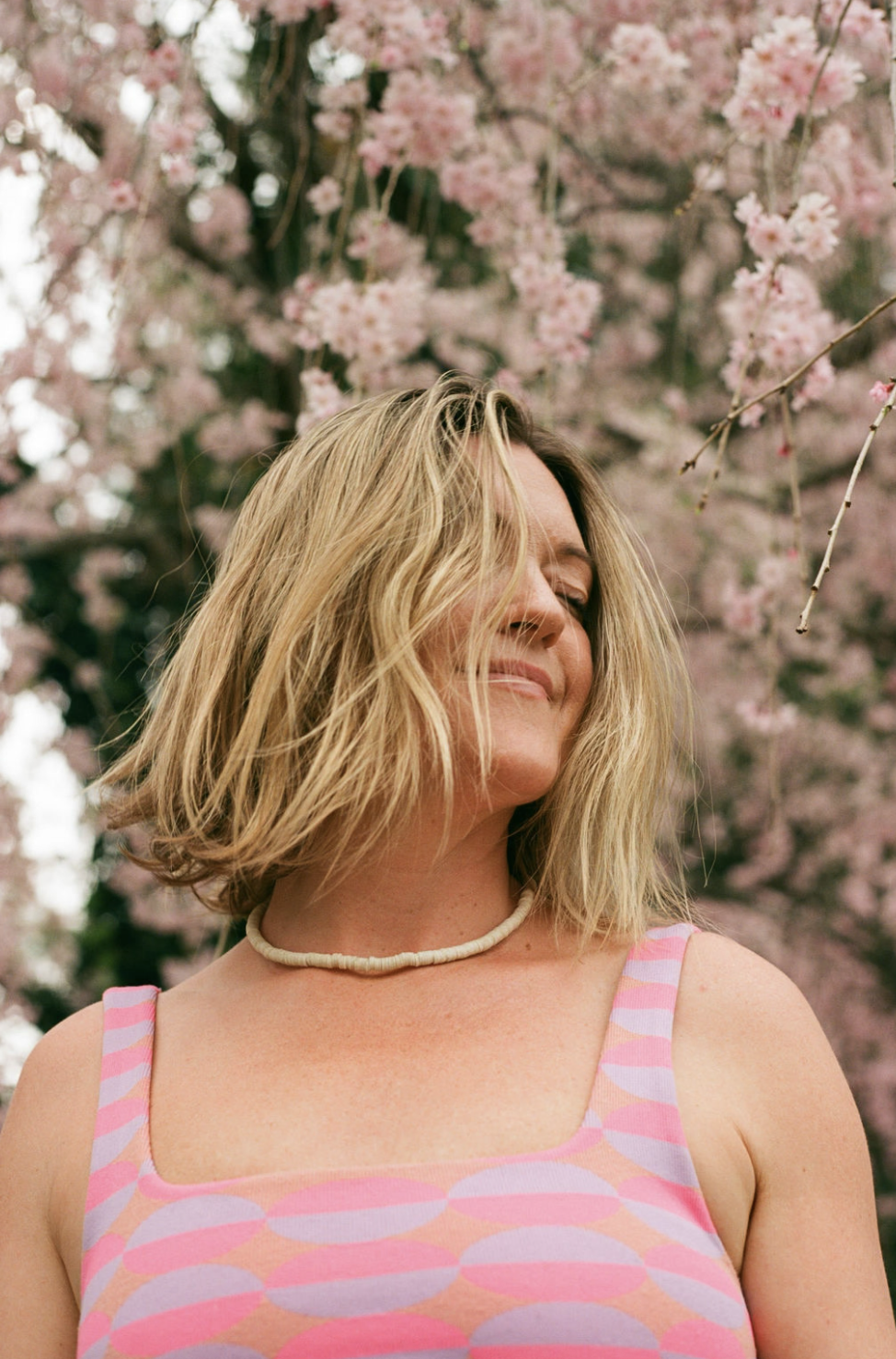 Woman with blonde hair smiling, standing in front of pink cherry blossoms, wearing a pink and purple patterned dress and a white beaded necklace.
