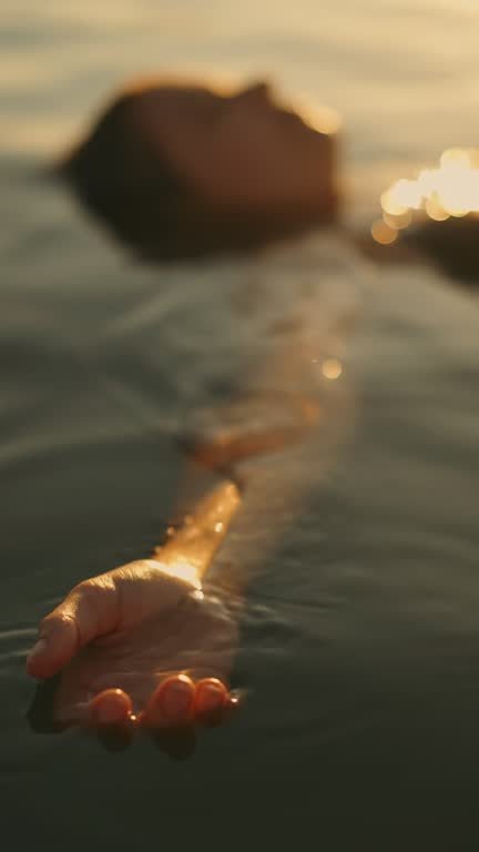 Close-up of a person’s hand reaching out from water with a blurred rock in the background during sunset.