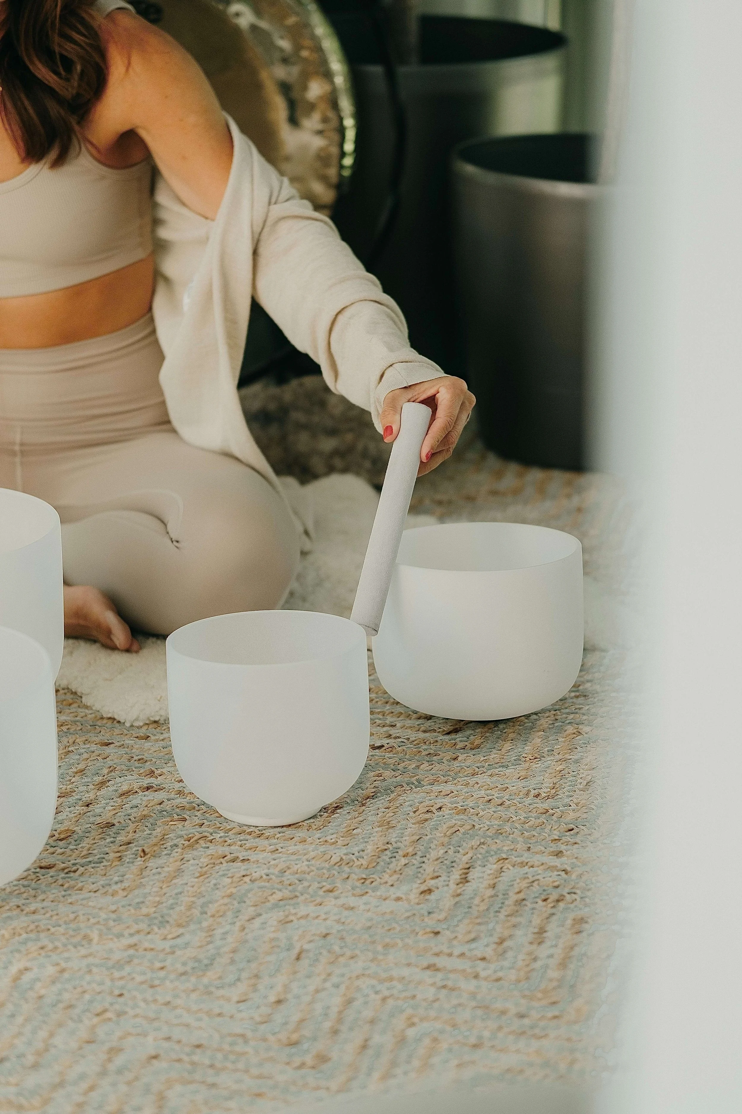 A person sitting on a patterned rug playing a set of white crystal singing bowls with a mallet in a cozy room.