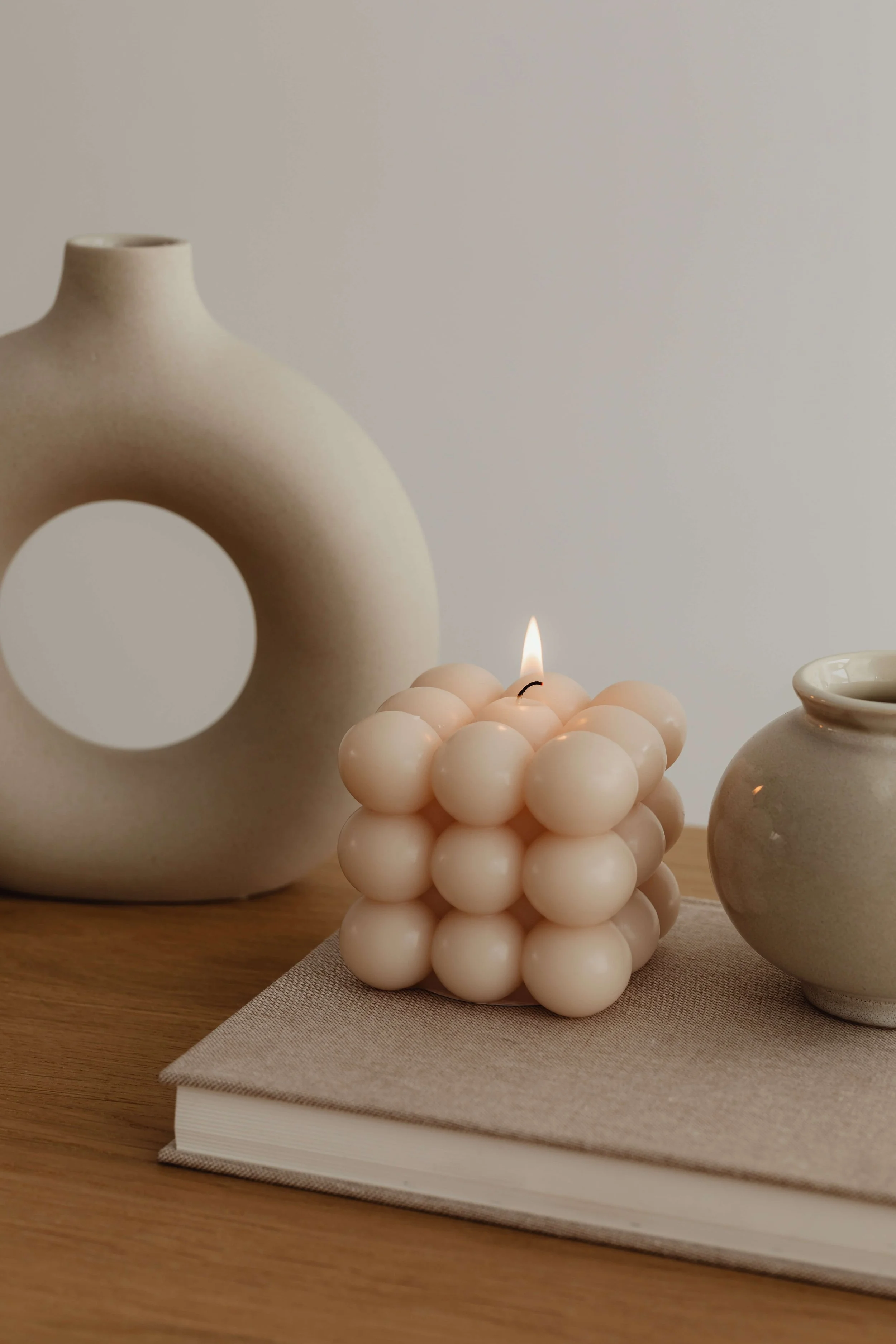 A table with a beige notebook, a candle made of interconnected round spheres with a small flame on top, a large minimalist ceramic vase with a hole in the middle, and a smaller ceramic container, all placed against a plain wall.