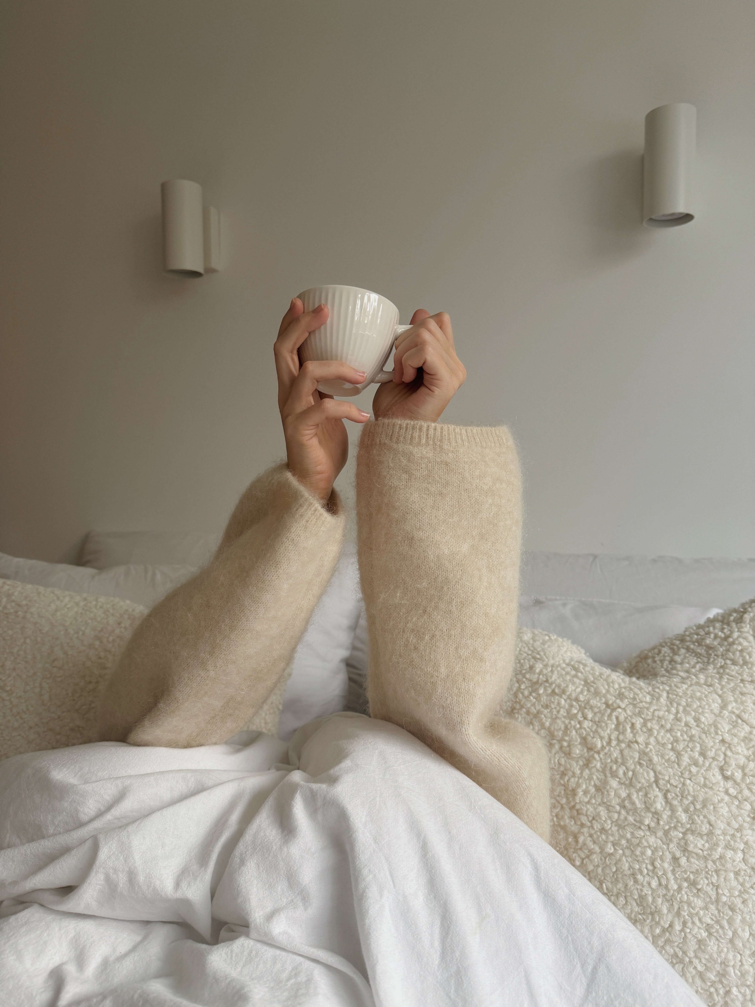 Person lying in bed, holding a white mug up towards the ceiling, wearing beige fuzzy socks and a cream sweater, with a white bedsheet and pillows in a neutral-colored bedroom.