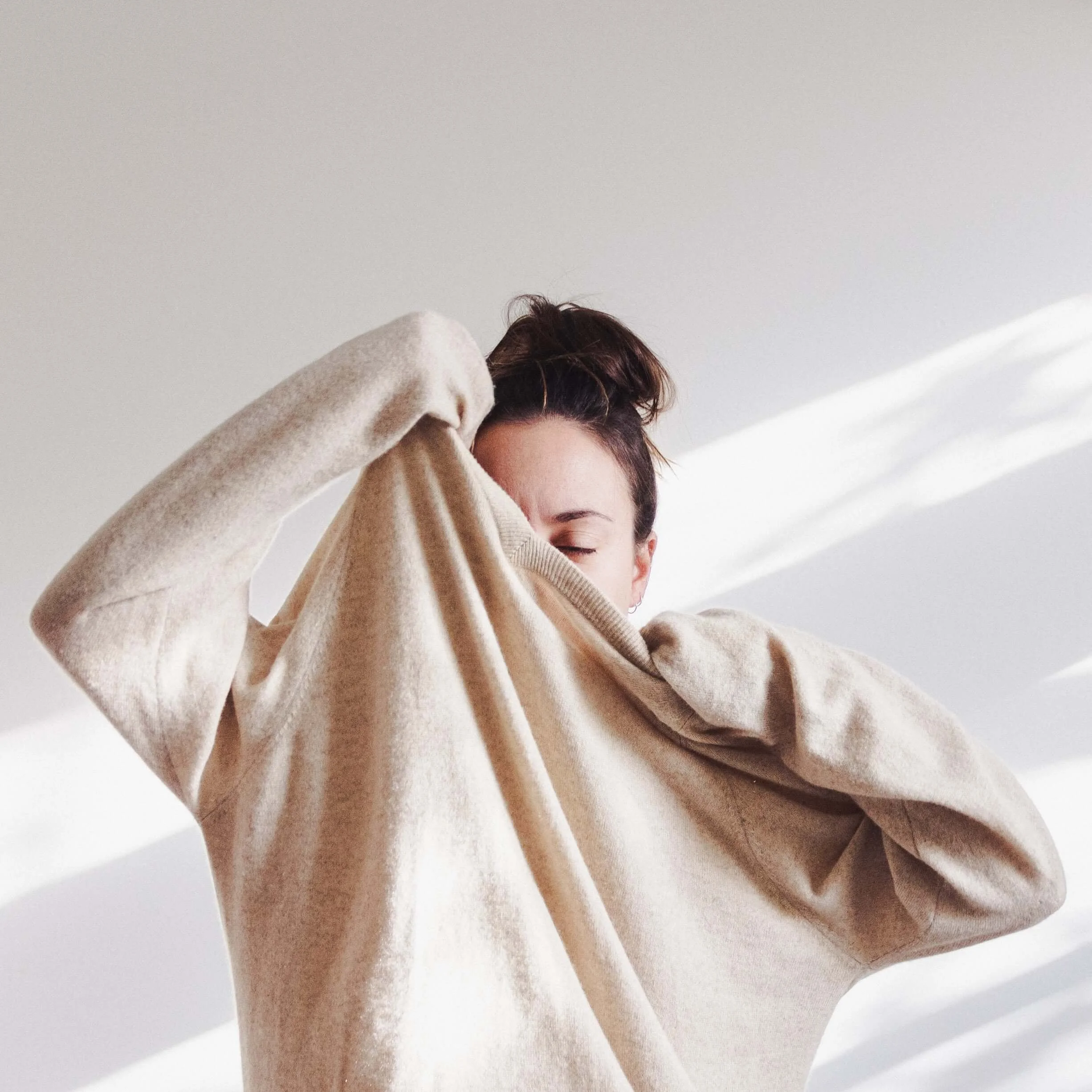 A woman with brown hair in a bun is lifting the collar of her beige sweatshirt over her face, with her eyes closed, against a white background.