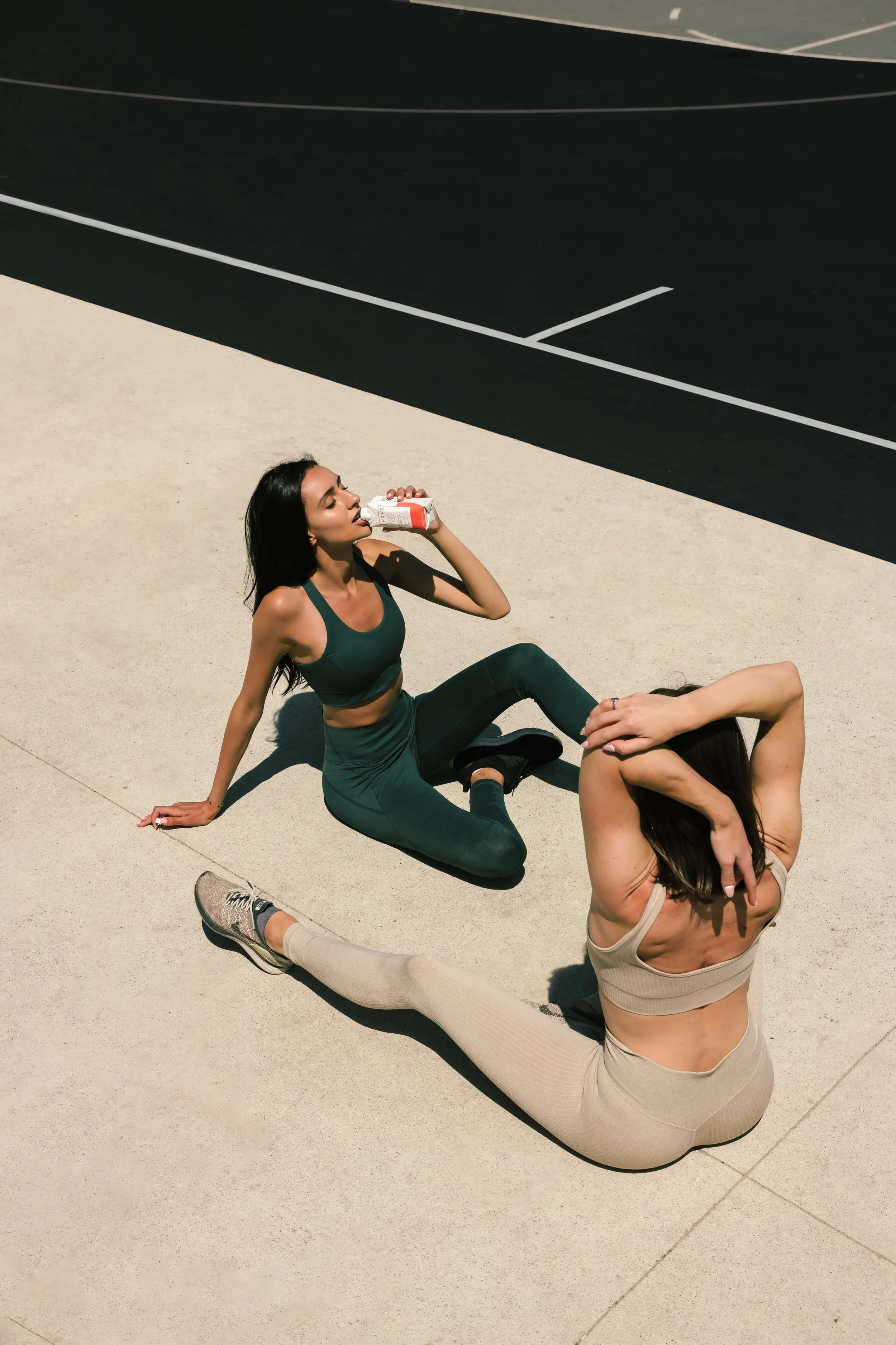 Two women sitting on the ground outdoors, one drinking from a small bottle, the other stretching her arm over her head.