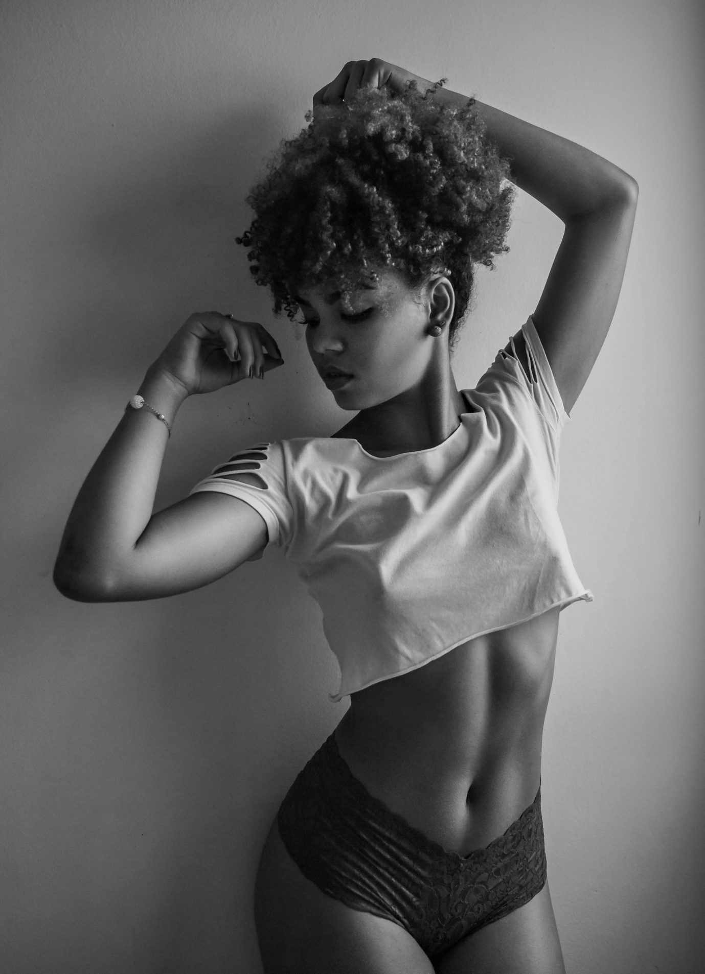 A woman with curly hair poses against a plain wall, wearing a cropped t-shirt and lace underwear.