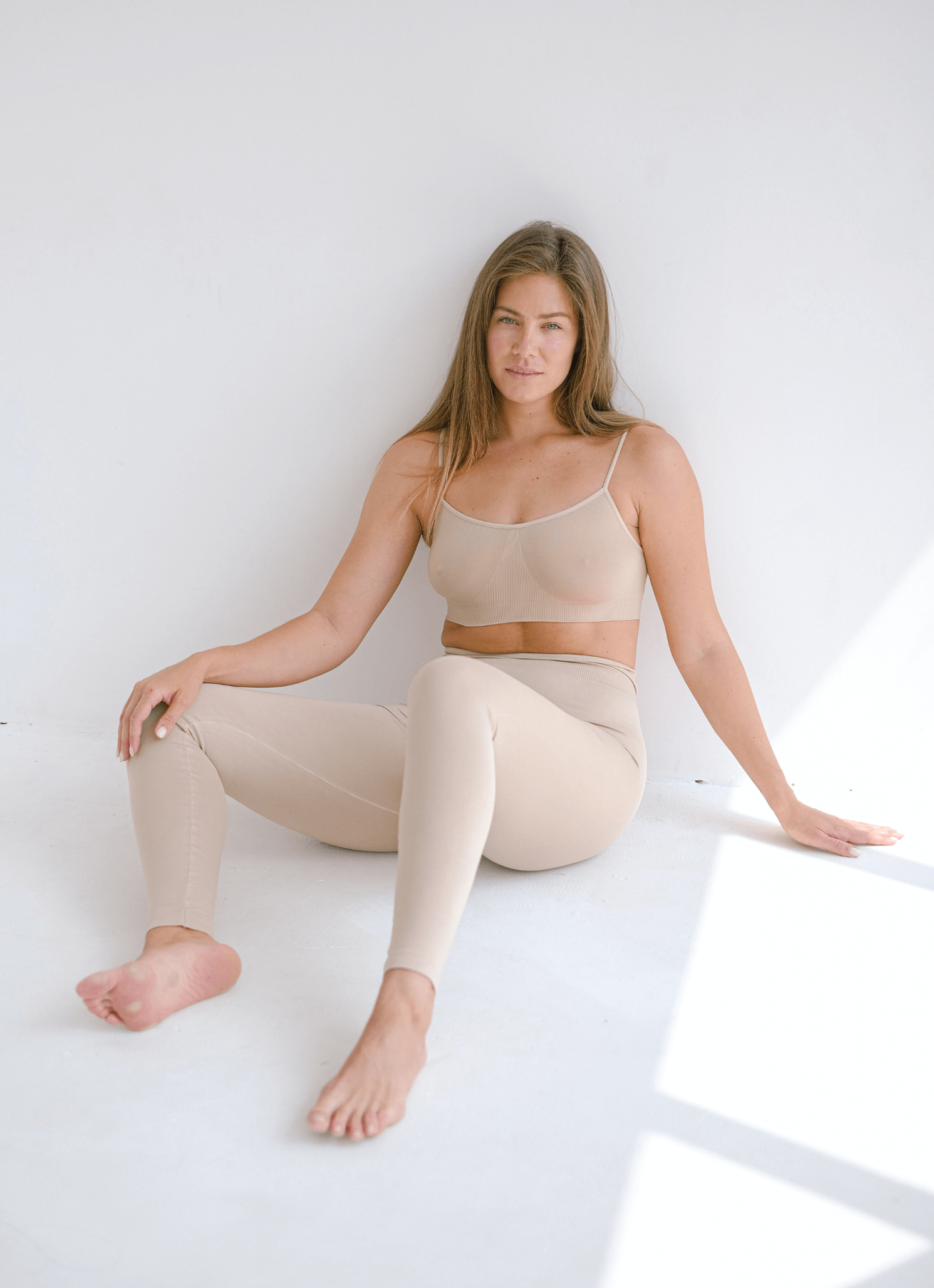 A woman with light skin and long brown hair sits on the floor in a white room, wearing beige leggings and a beige crop top.